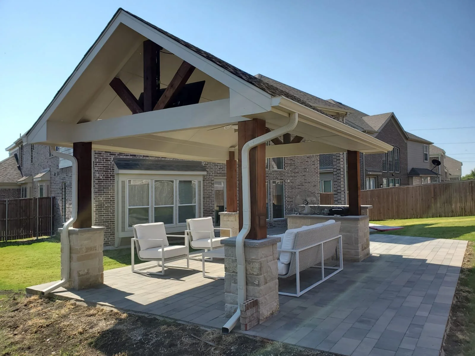 Covered backyard patio (with stone work for flooring) with outdoor furniture under the covered patio, brick house in the background, and a wooden fence.