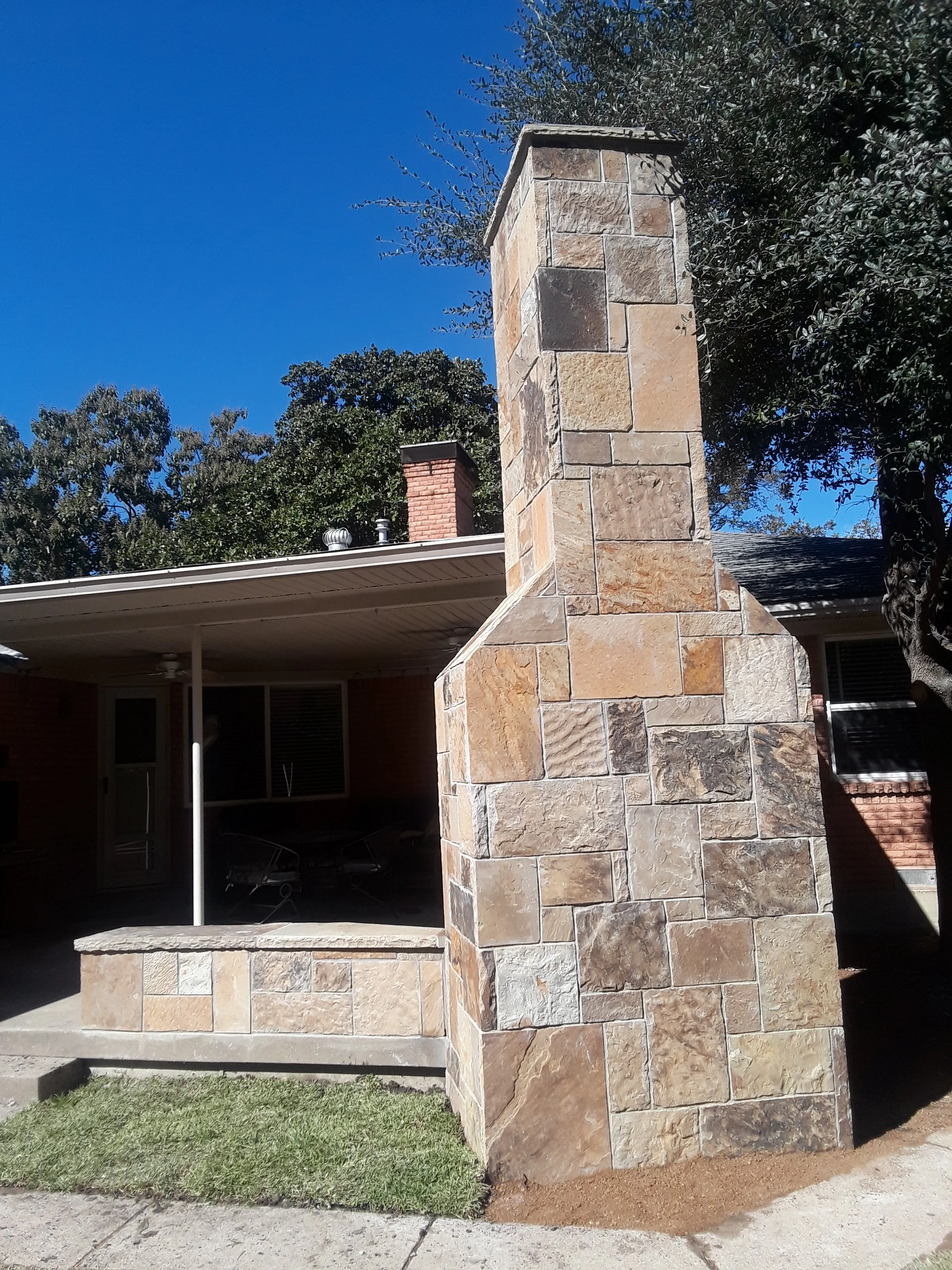 A stone chimney on a house, with a covered patio area underneath. There is a green lawn and trees in the background under a clear blue sky.
