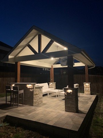 A modern outdoor patio area with a roof, seating, and lighting, illuminated at night.