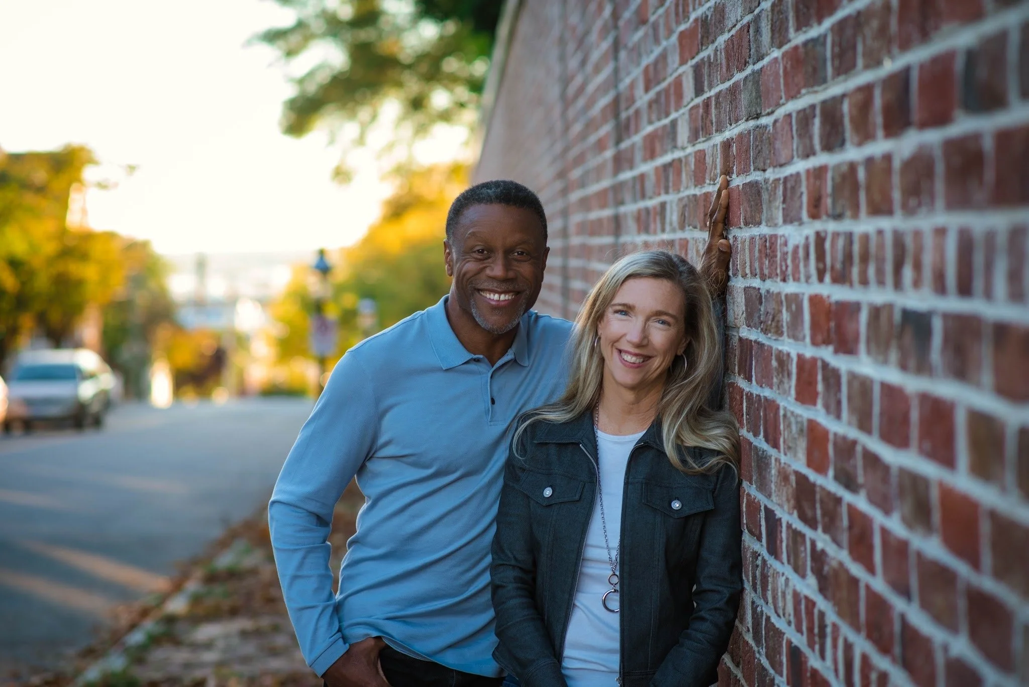 A smiling middle-aged African American man and a Caucasian woman leaning against a brick wall outdoors during daytime, with trees and a street in the background.