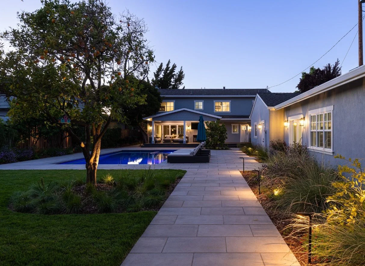 Backyard with pool, patio area, and house, illuminated at dusk, with tree, lounge chairs, and garden lighting.