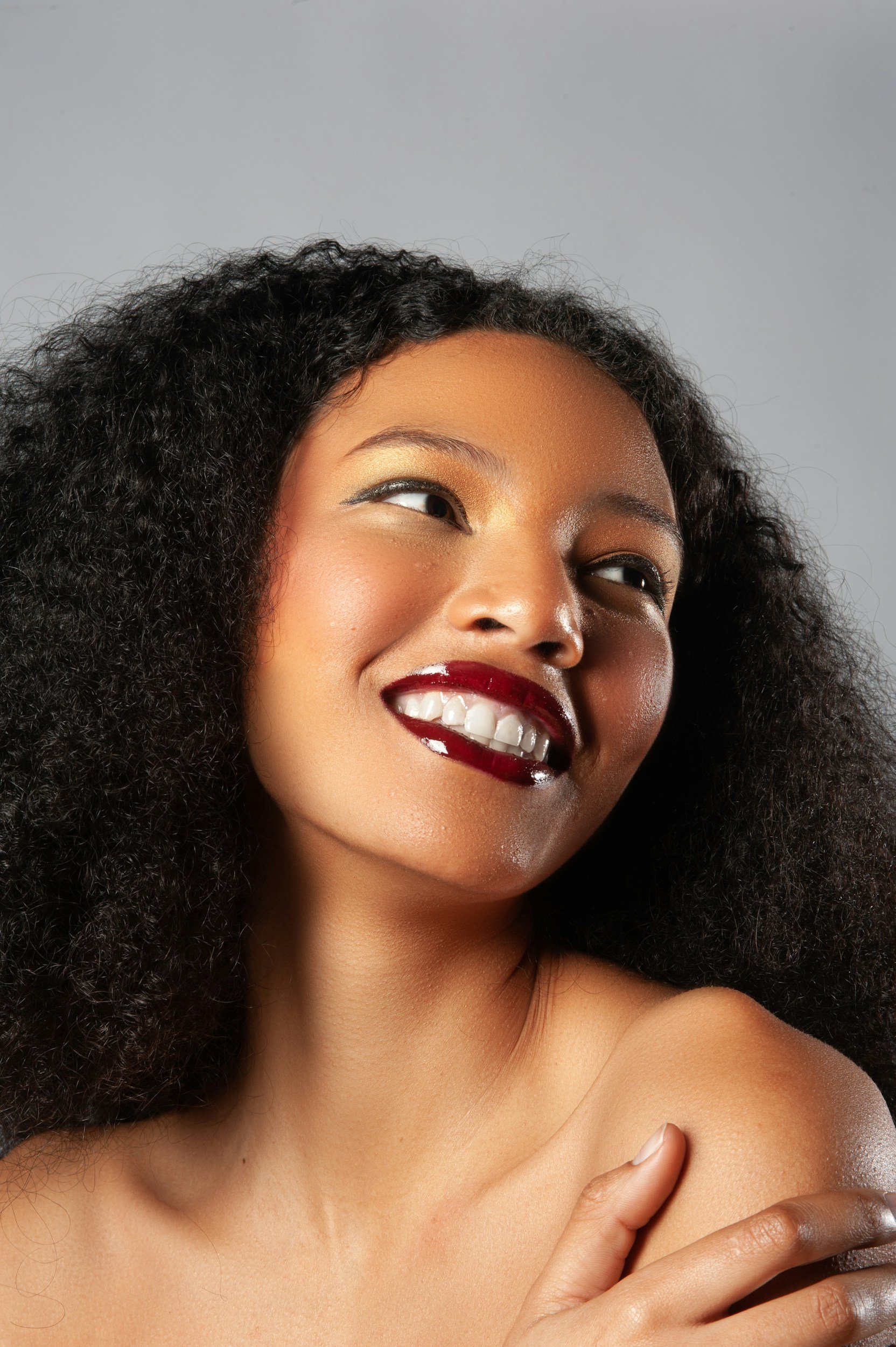 Close-up of a smiling woman with dark curly hair, wearing makeup, and touching her shoulder.