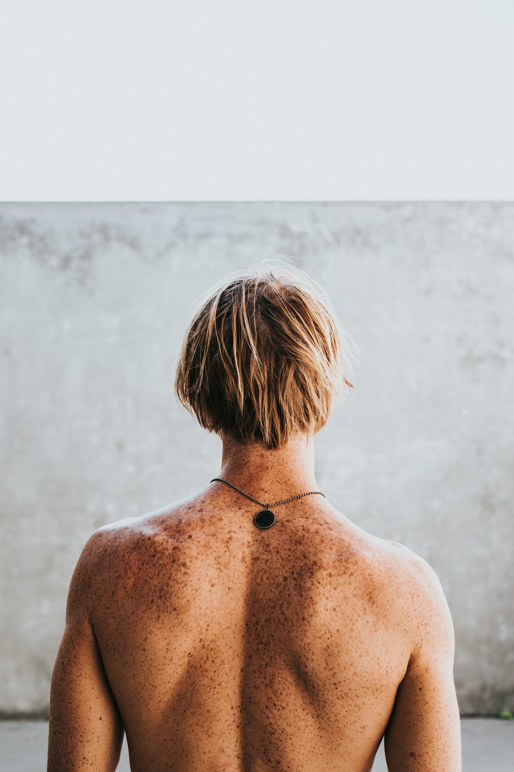 Back of a person with short, light brown hair and freckles, wearing a necklace with a circular pendant, standing topless against a plain, light-colored wall.
