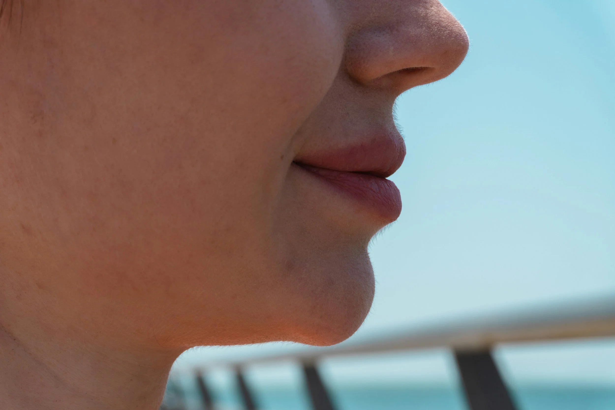 Close-up of a person's profile, focusing on the nose, lips, and part of the cheek, with a light blue sky and blurred wooden structure in the background.