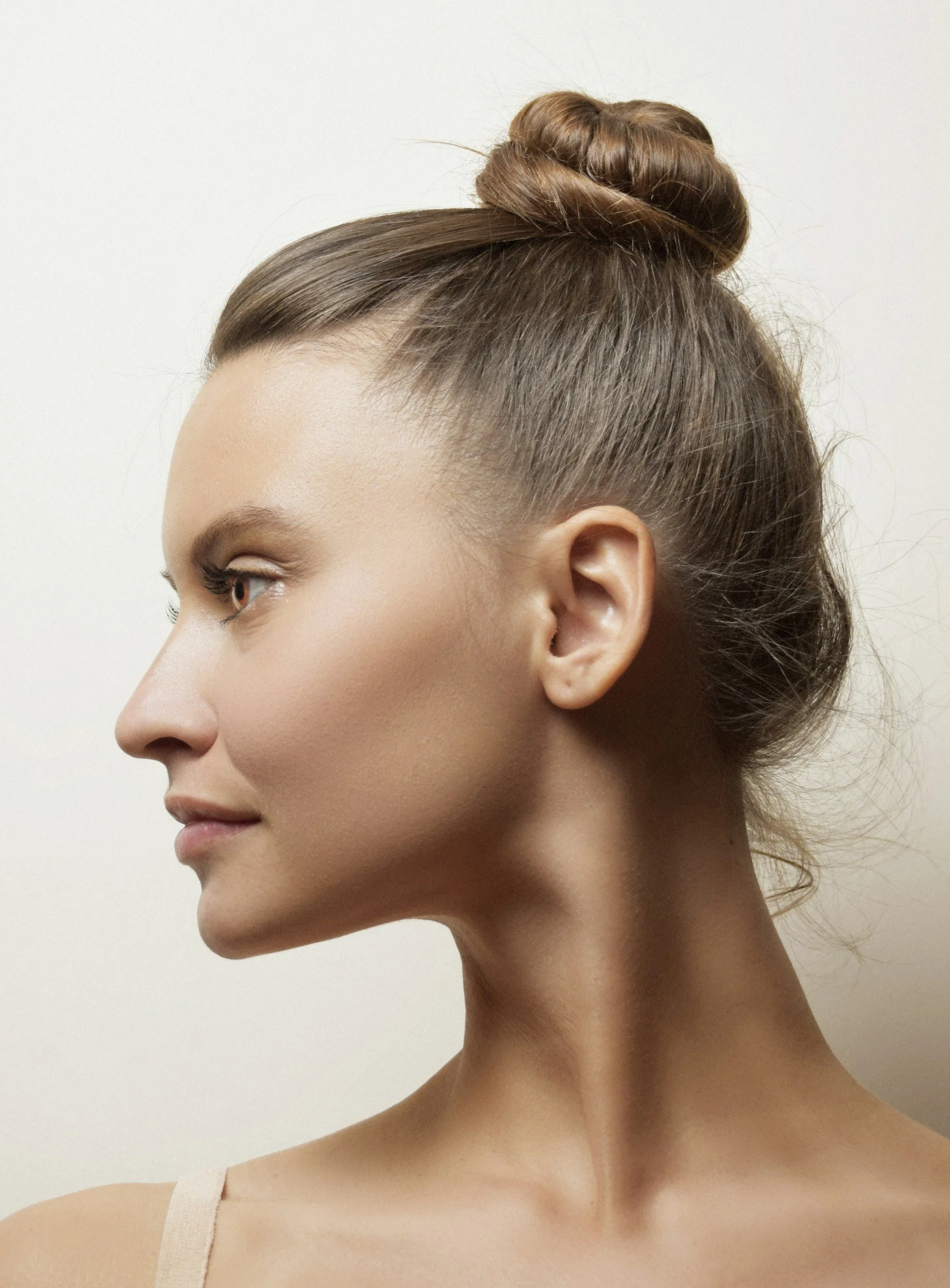Profile of a young woman with styled brown hair in a bun, wearing a beige tank top, against a plain white background.
