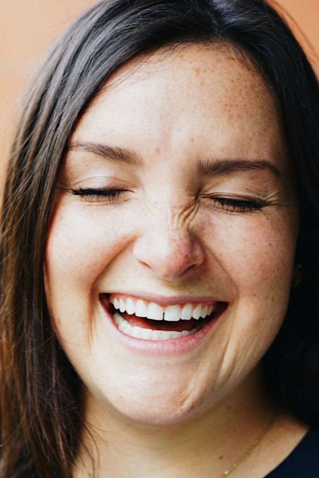 A woman with dark hair smiling and laughing with eyes closed, showing her teeth.