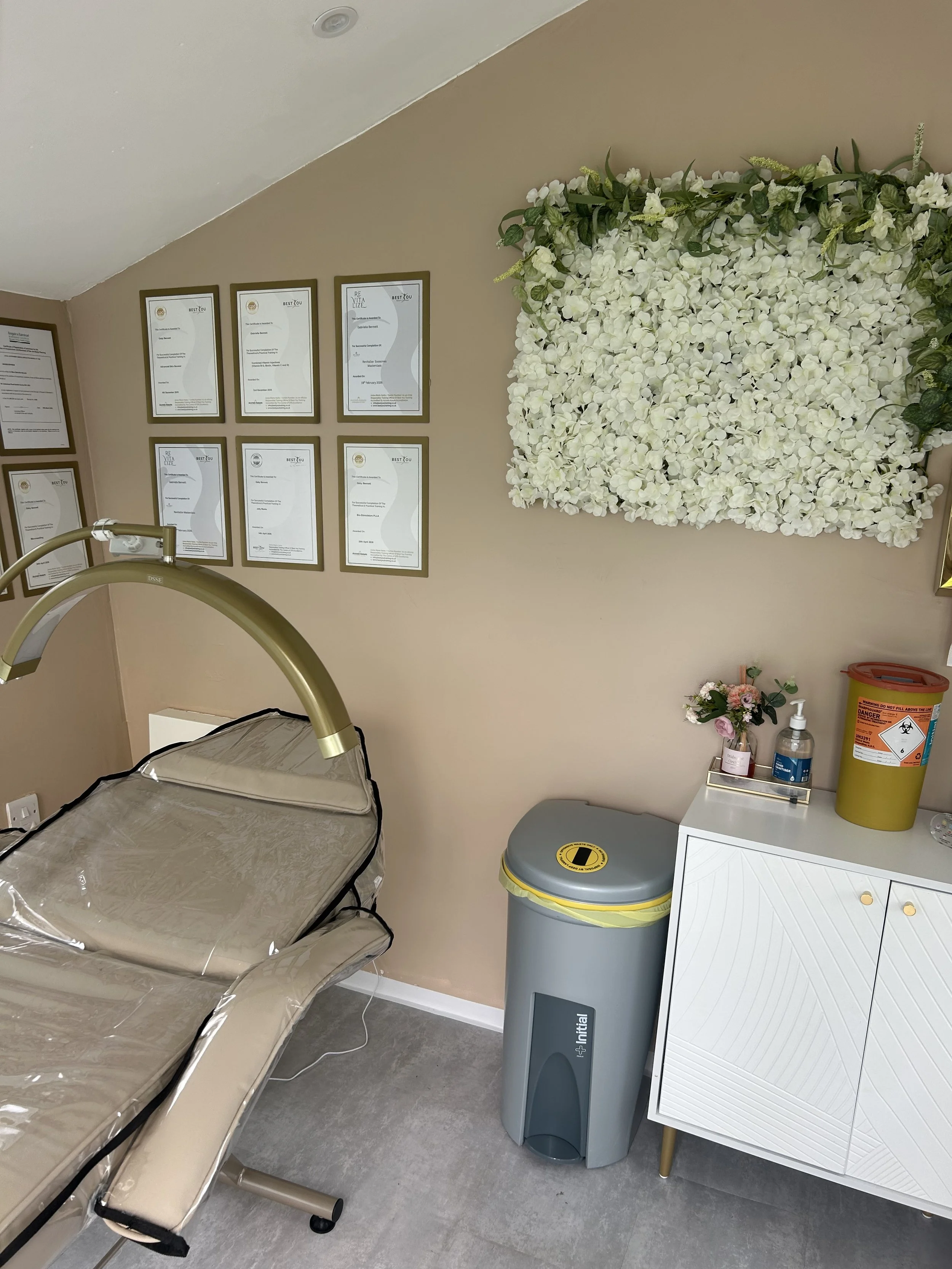 Hospital or medical clinic room with a beige wall, framed certificates or diplomas, a large floral wall decor, a small table with a pink flower arrangement, hand sanitizer, a yellow caution bin, a medical examination bed, and medical equipment.