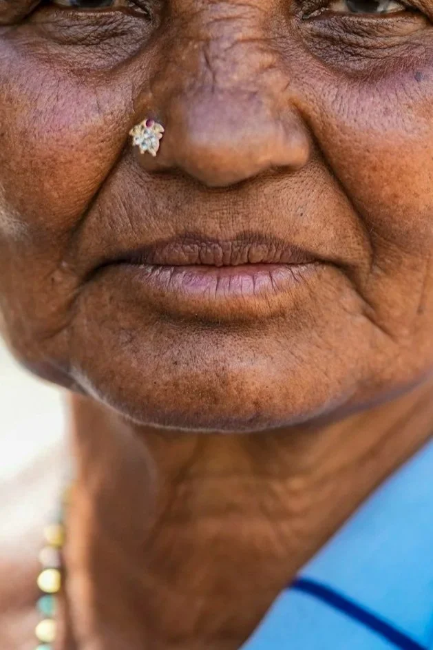 Close-up of an elderly person's face, showing a nose piercing with a small flower-shaped jewel and wrinkled skin.