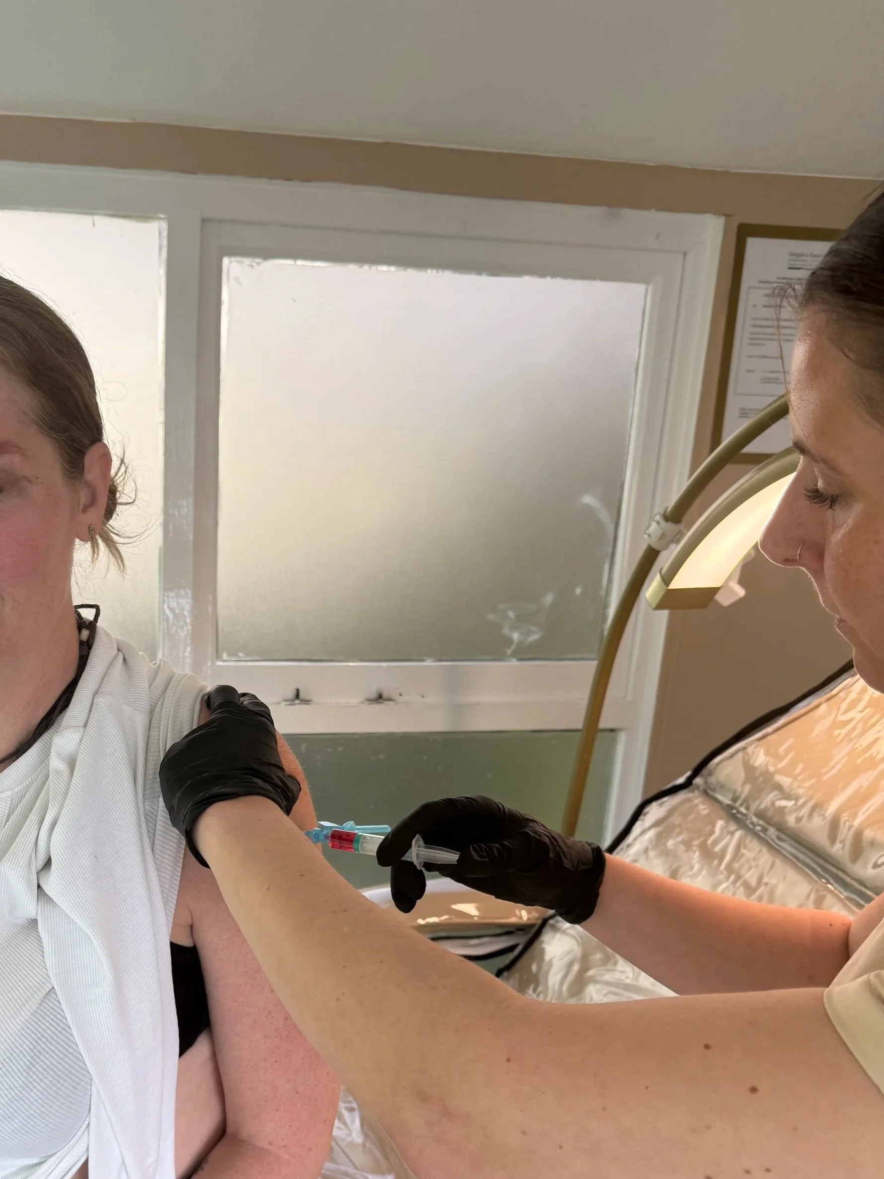 A person receiving a vaccination shot in their upper arm from a healthcare worker who is wearing gloves and protective gear, in an indoor medical setting.