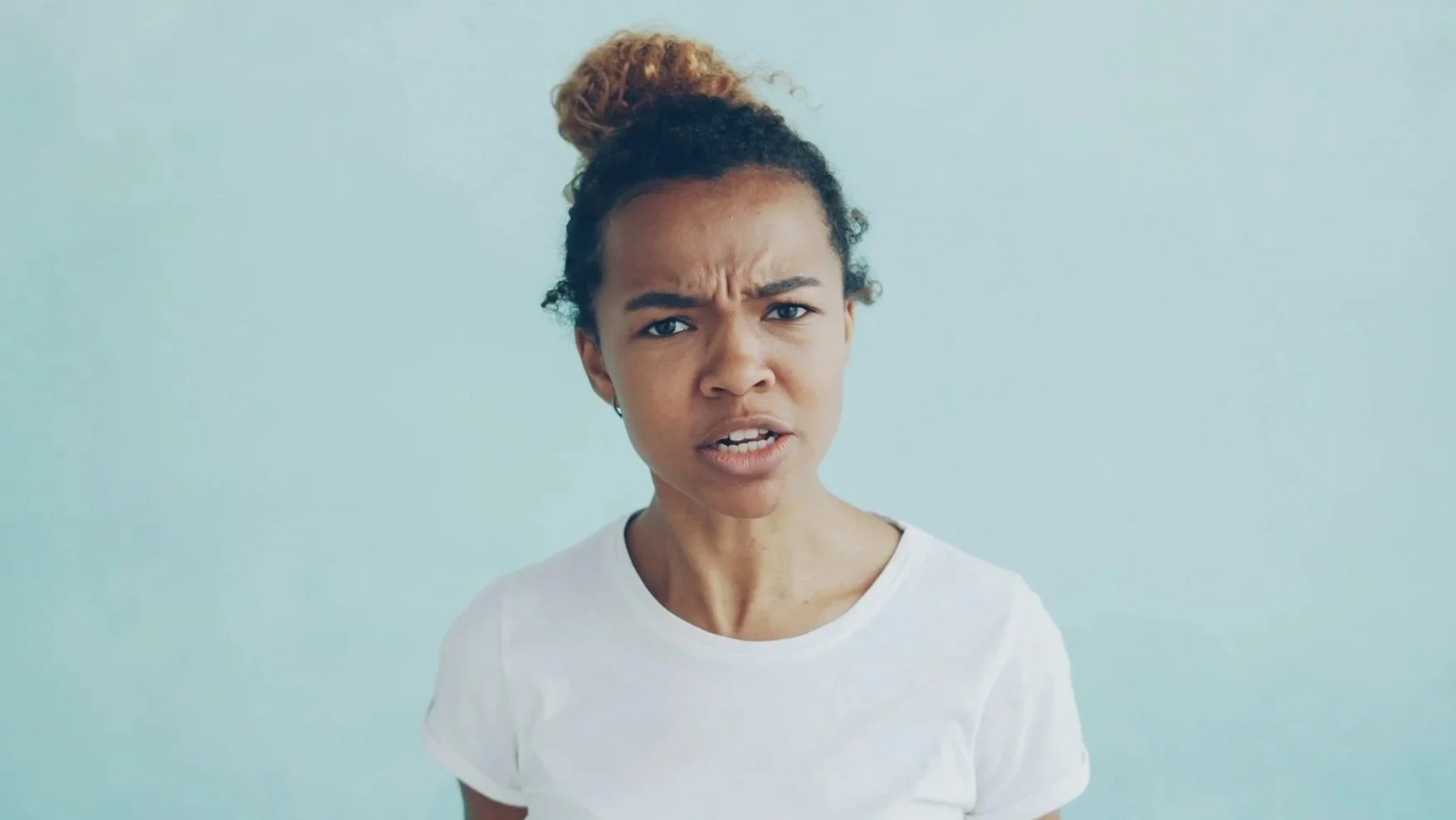 A woman with a stressed or angry expression, furrowing her eyebrows and slightly opening her mouth, standing against a plain light blue background.