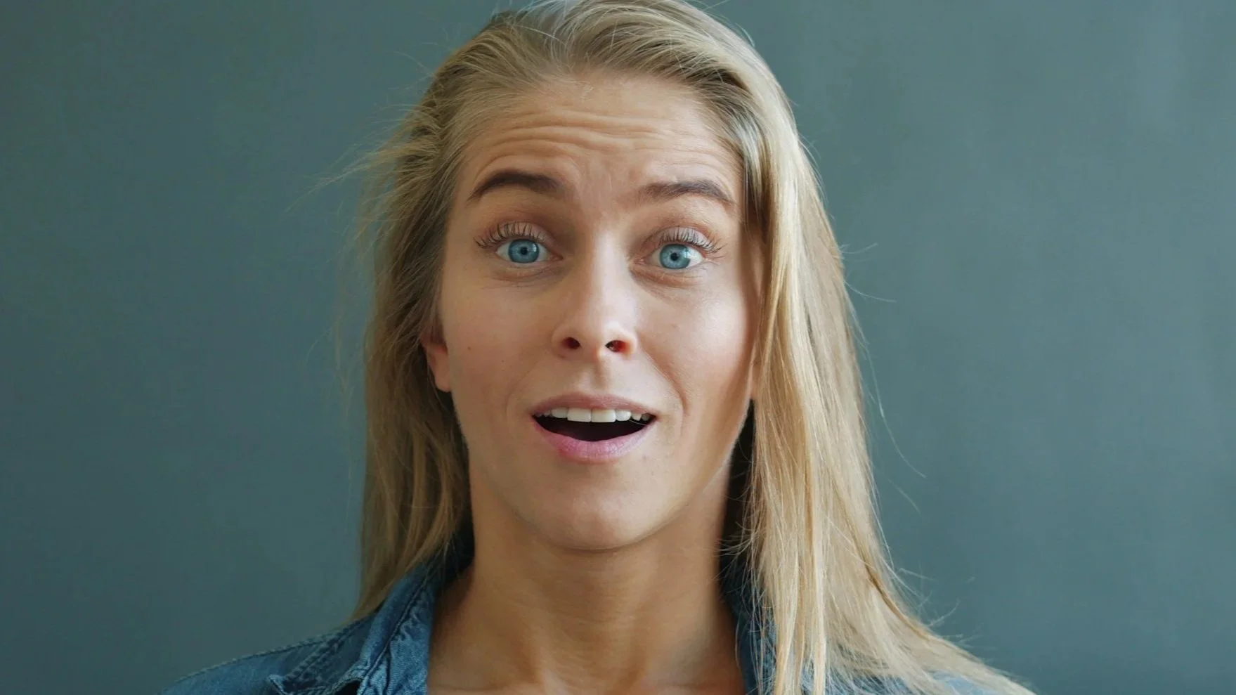 Close-up of a woman with long blonde hair, wide blue eyes, and an open-mouthed surprised expression, wearing a denim jacket against a gray background.