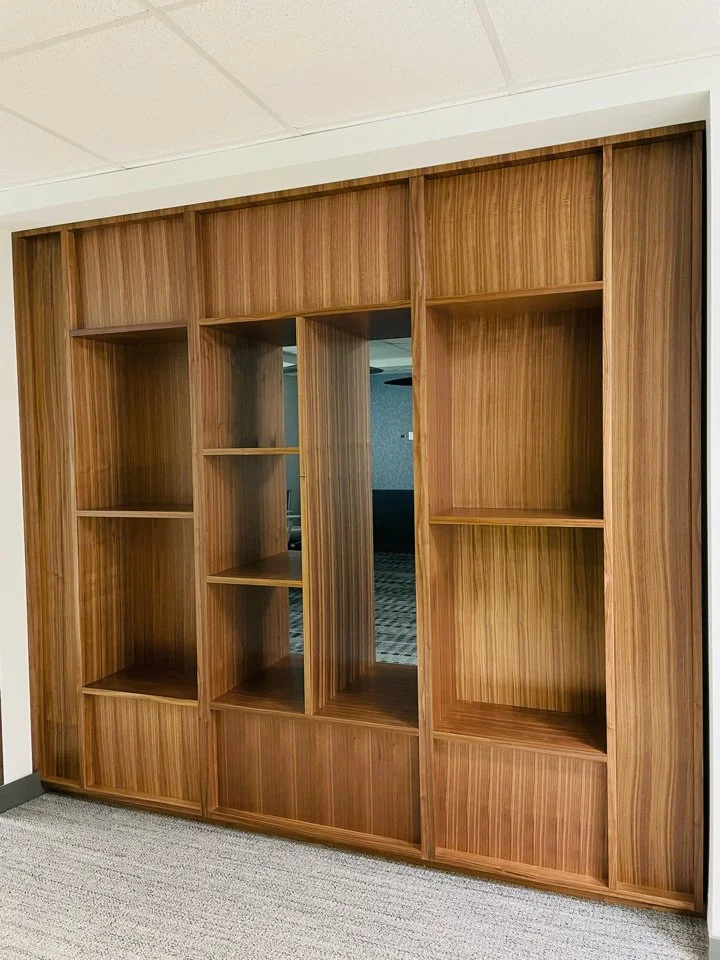 Empty wooden bookshelf with multiple shelves, positioned against a white wall in a room with carpeted floor.