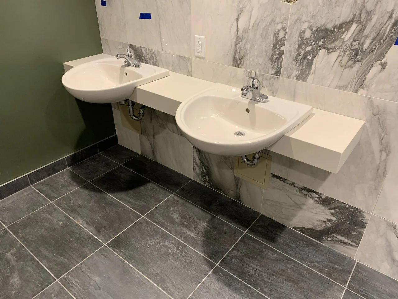 Two white sinks with chrome faucets mounted on a beige floating countertop in a bathroom with marble wall tiles and dark gray floor tiles.