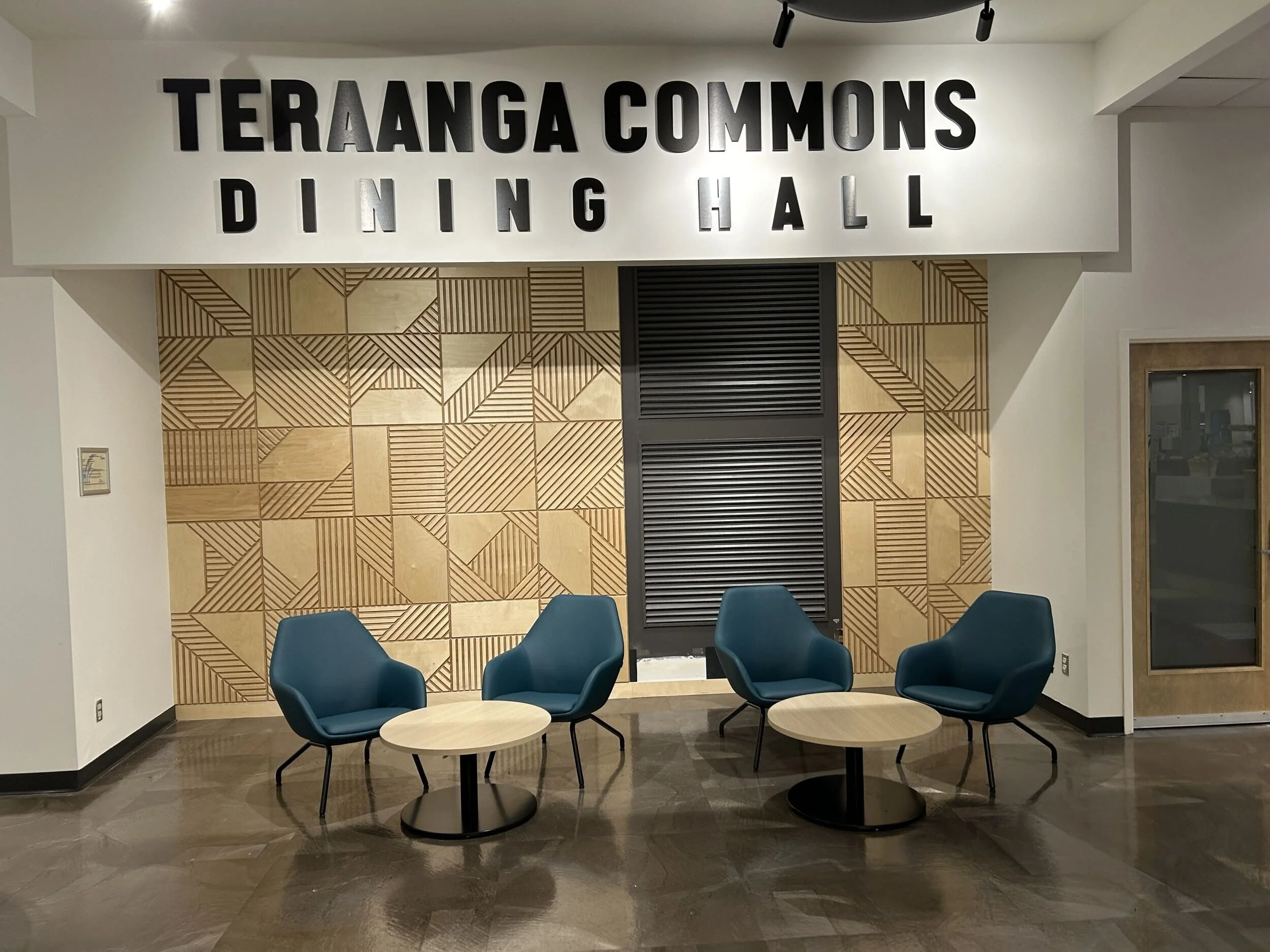 A waiting area in a dining hall with four blue chairs and two round wooden tables in front of a decorative wall with geometric wooden patterns and black and silver text on the wall that says, 'TERANGA COMMONS DINING HALL'.