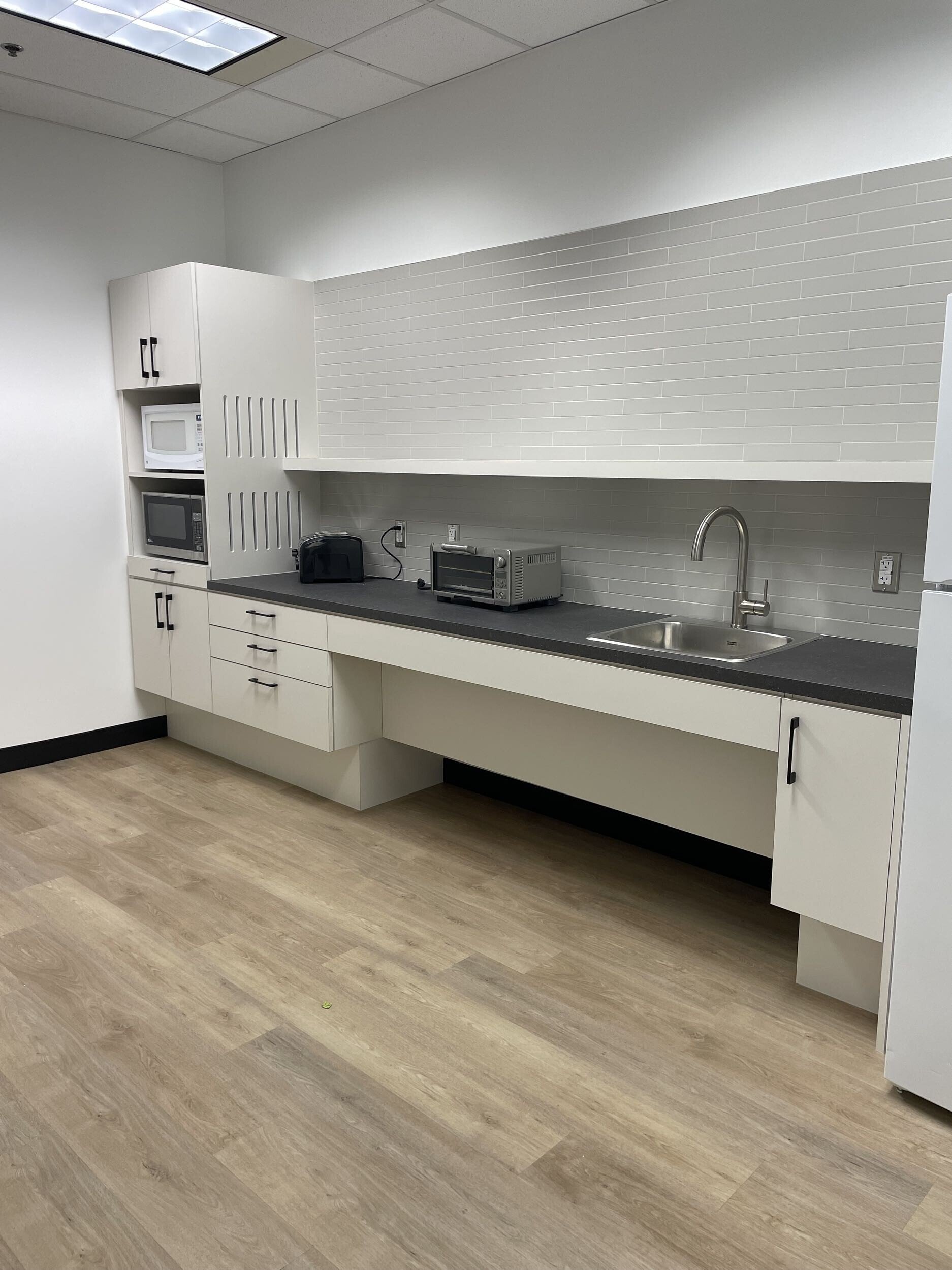 A minimalist kitchen with light wood flooring, white cabinetry, a black countertop, and a white tiled backsplash. Contains small appliances like a microwave, toaster, and oven, along with a stainless steel sink.