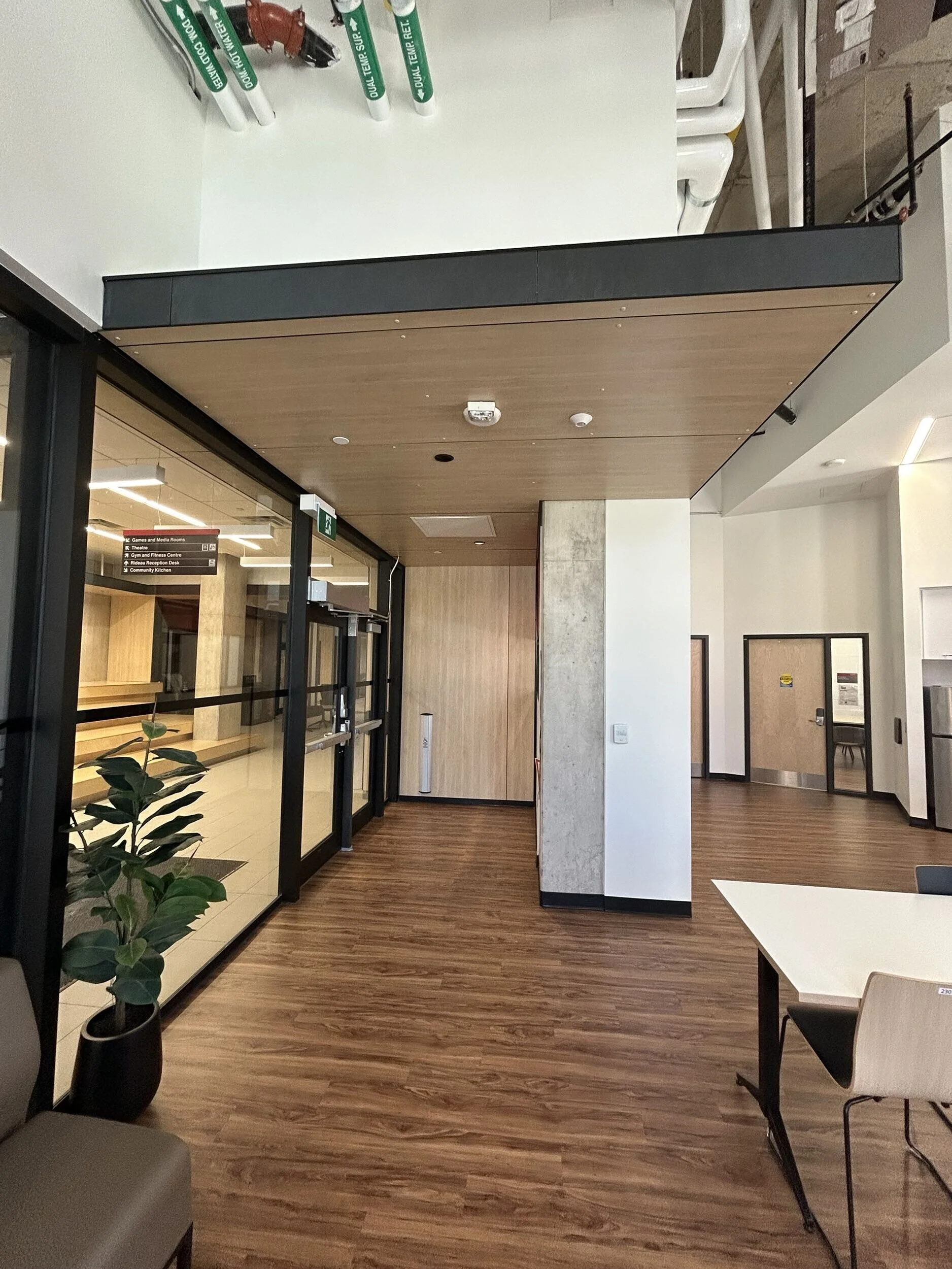Interior view of a modern lobby or waiting area with wood flooring, a potted plant, glass walls, and a reception desk visible in the background.