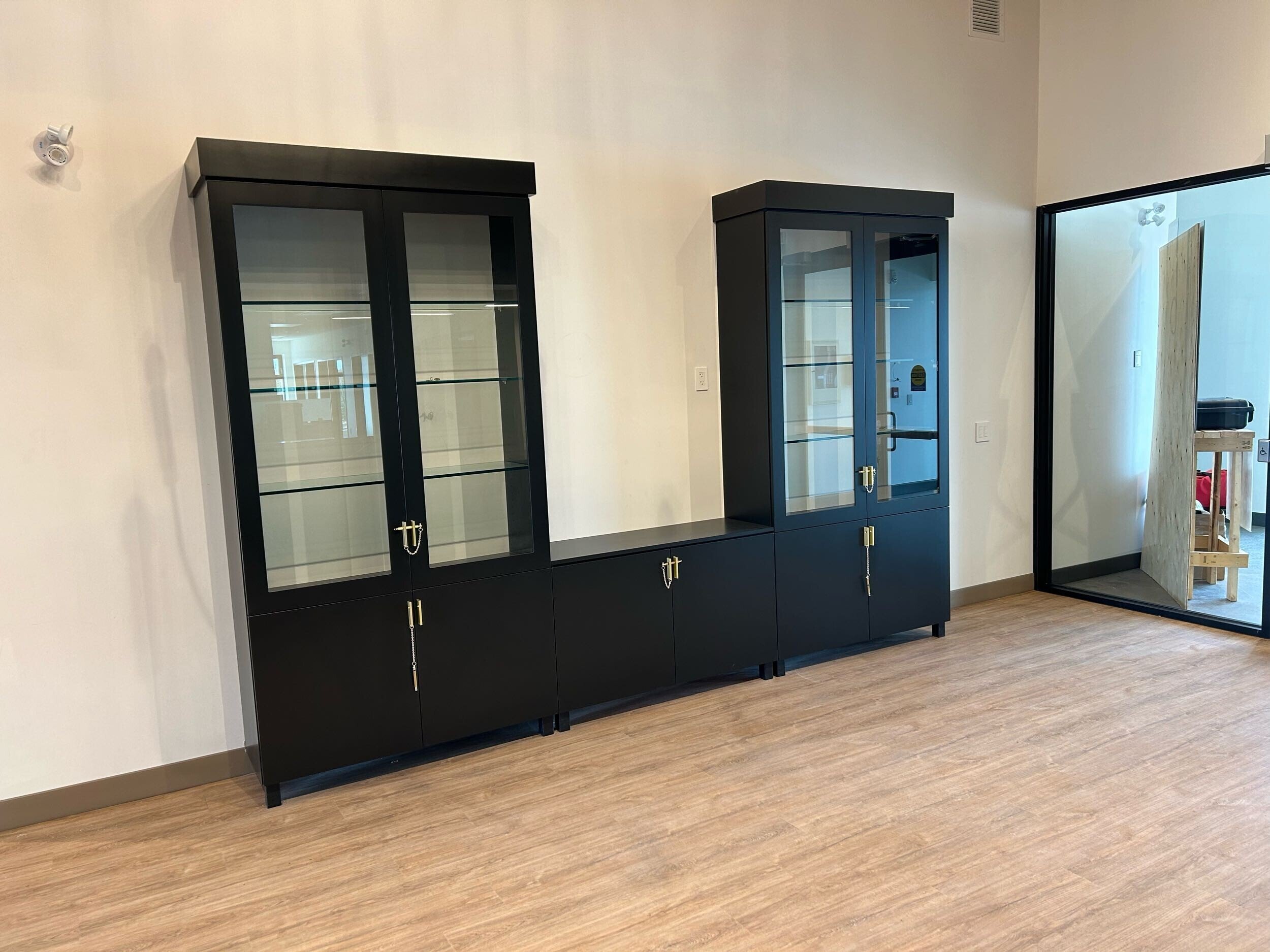 Black display cabinet with glass doors and gold handles standing against a white wall in a room with wooden floor.