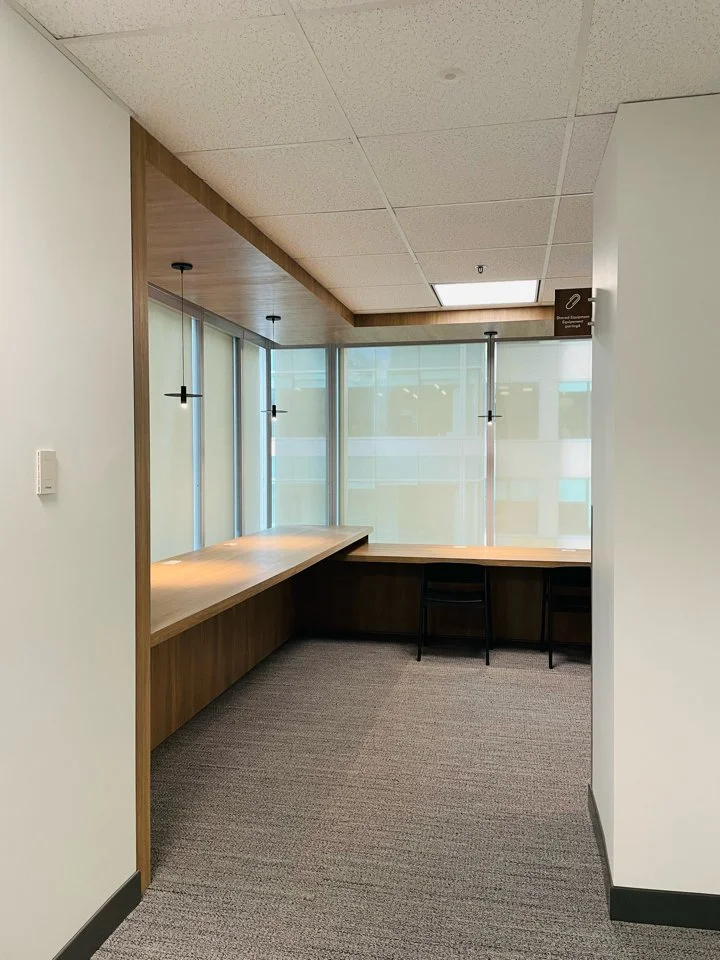 Corner office with large windows, a long wooden desk, two black chairs, and modern hanging pendant lights.