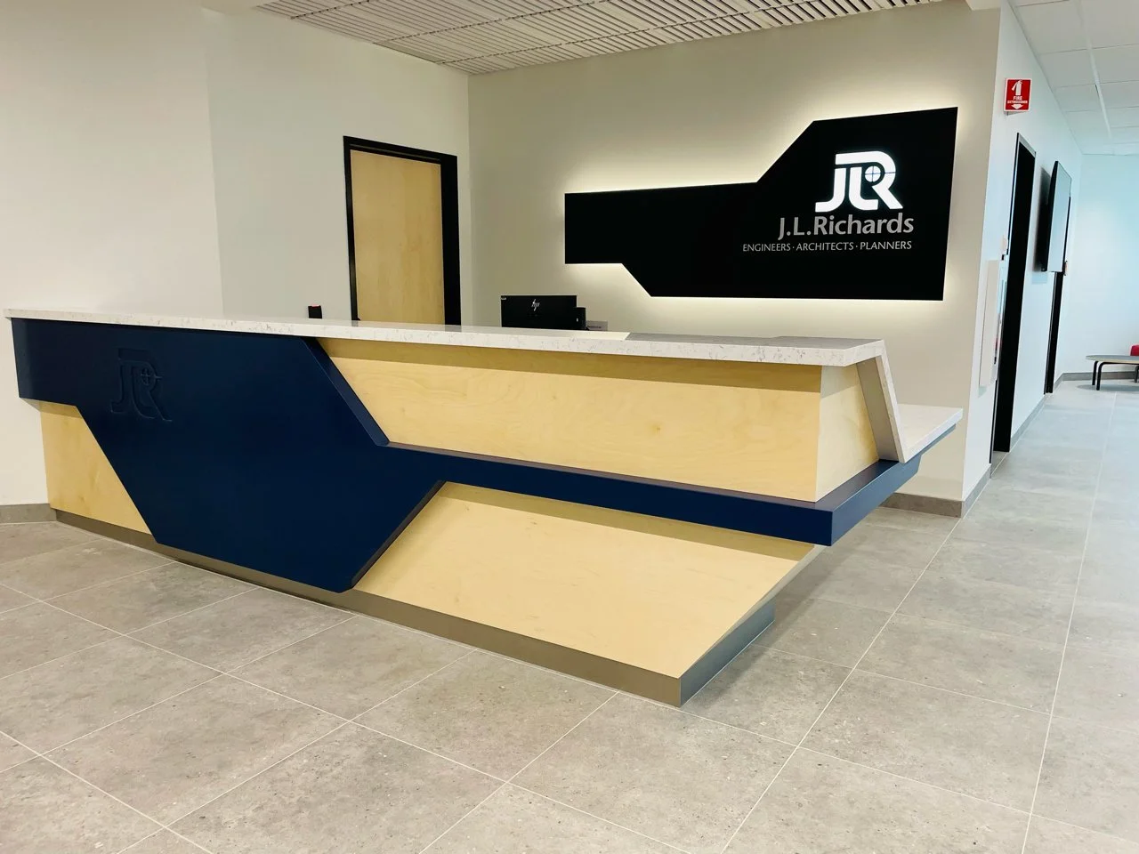 Modern reception desk with wood and dark blue accents in an office lobby, J.L. Richards logo on the wall, and a hallway with seating.