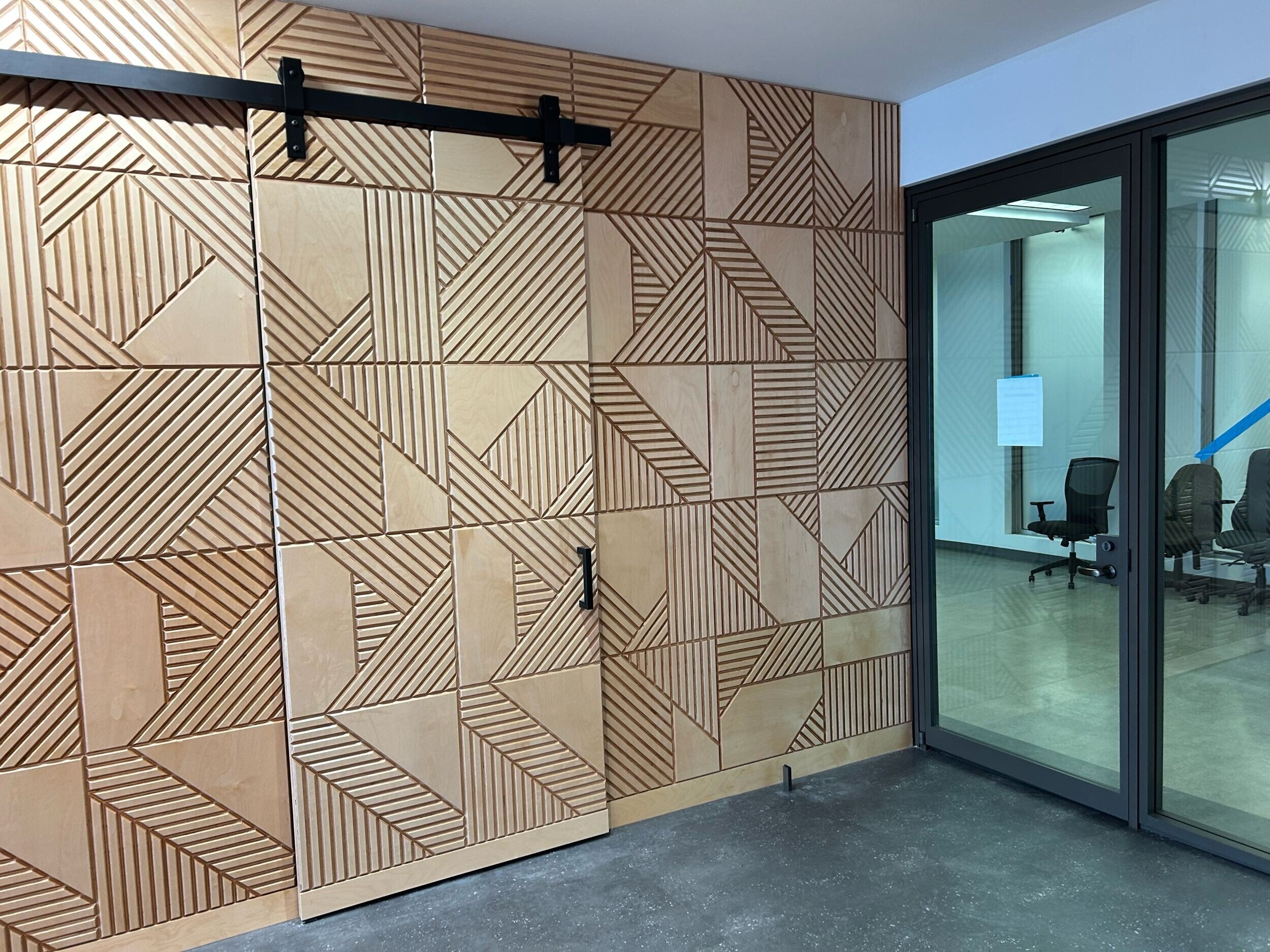 Interior office space featuring a wall with modern wooden geometric paneling design and a glass door to a conference room with office chairs inside.