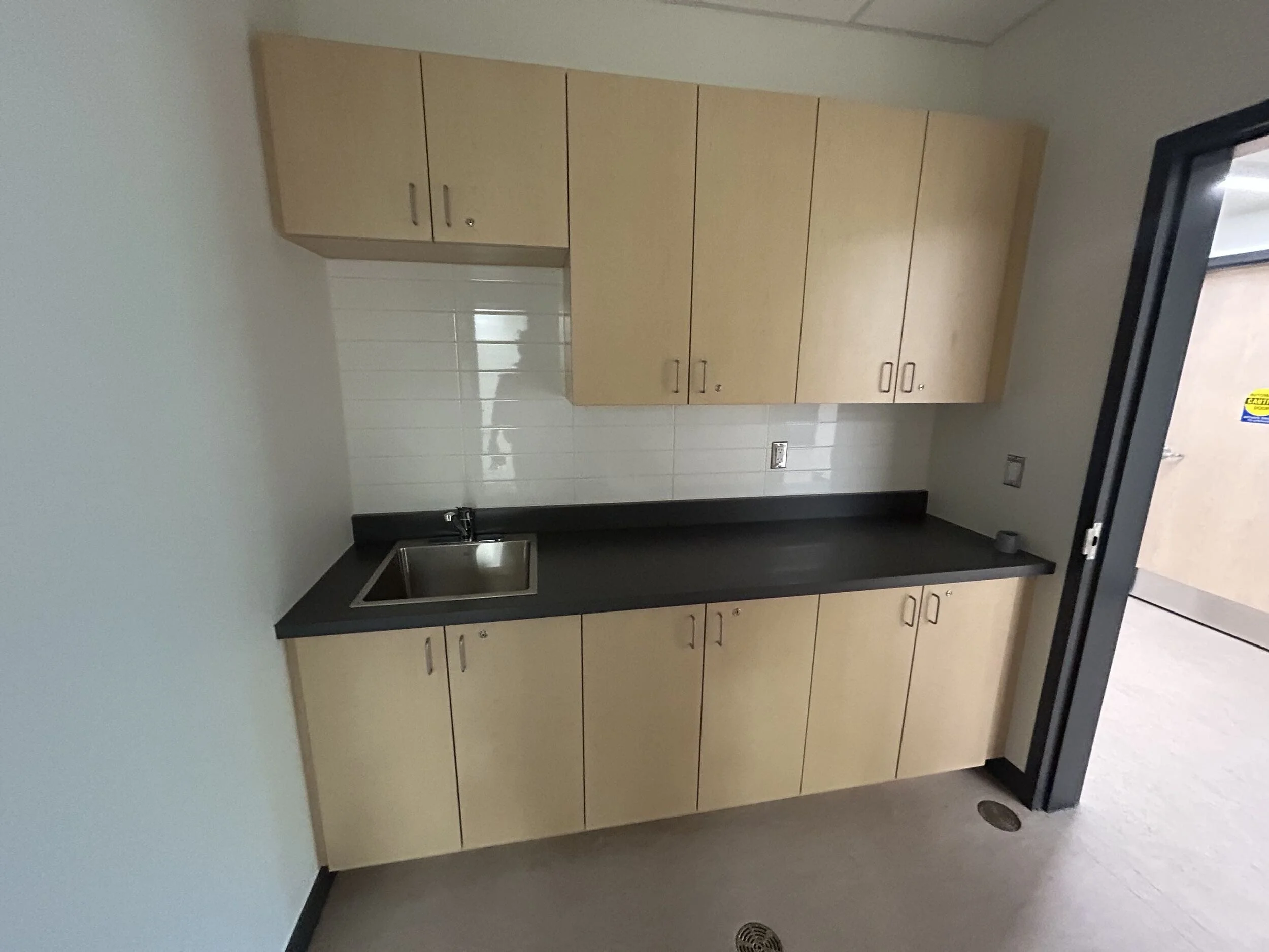 Empty room with a beige cabinet, a small stainless steel sink with a black countertop, white tiled wall, and a gray floor with a floor drain near the wall.