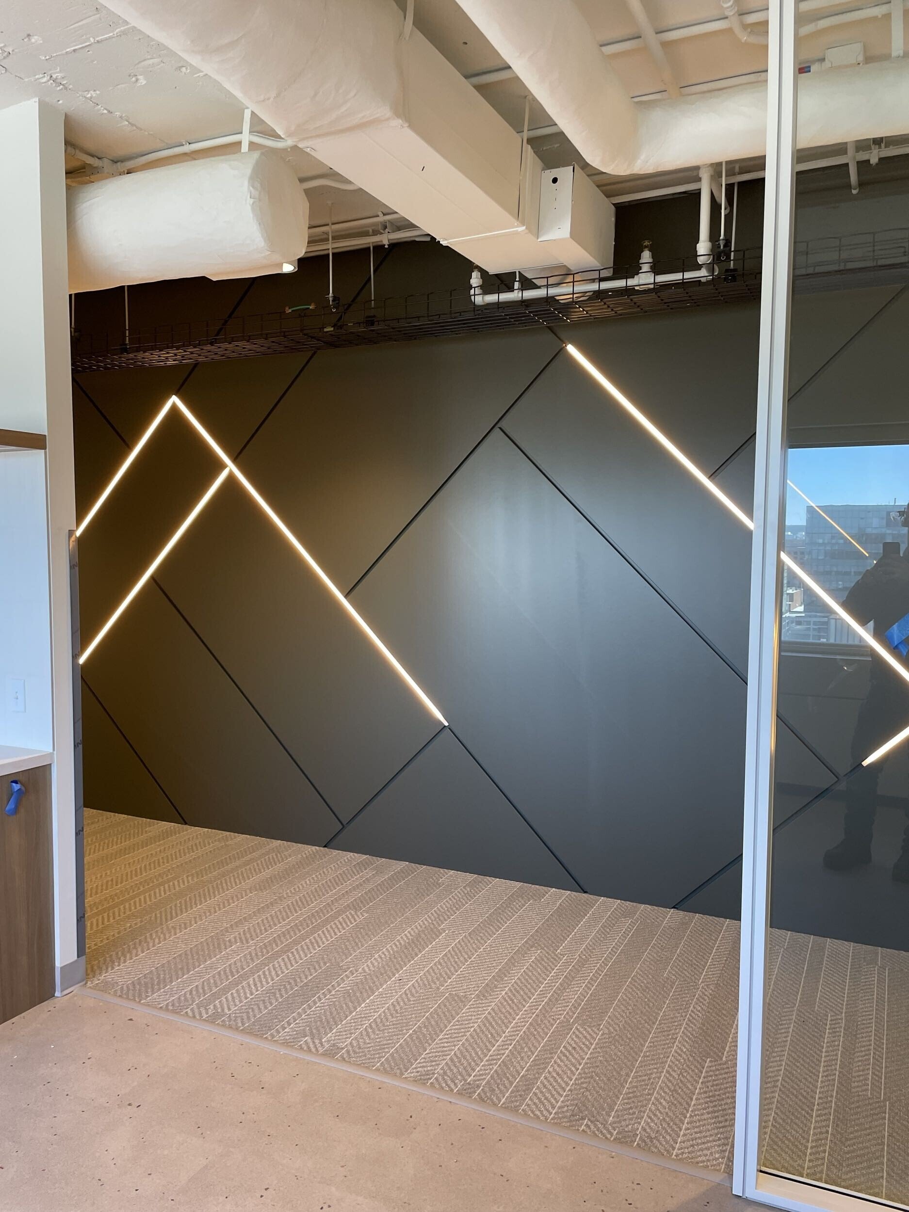Interior view of a modern building featuring a geometric black wall with diagonal lines and integrated linear lighting, a glass sliding door, and a curved beige carpeted floor.