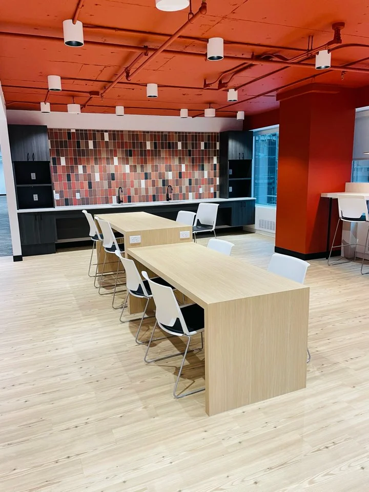 A modern meeting or conference room with a light wooden table and six white chairs. The room features a red ceiling with multiple white ceiling lights, a colorful mosaic tile backsplash behind a sink area, and large windows allowing natural light. Th