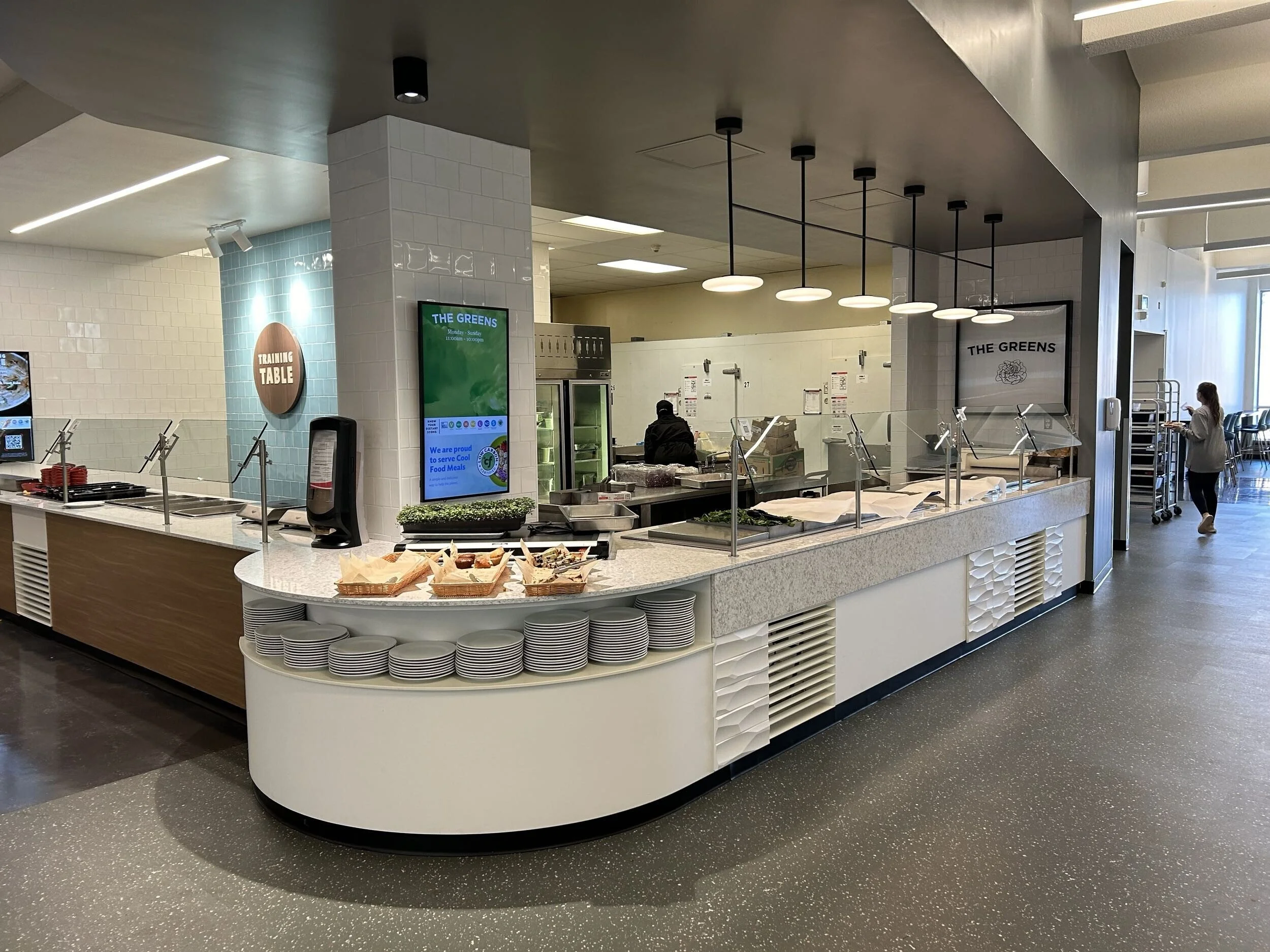 Food serving station with plates on display, digital menu screens, and a sign labeled 'Training Table' in a modern cafeteria setting.