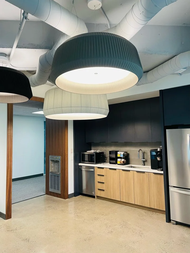 Modern office kitchen with large black and white pendant lights hanging from the ceiling, featuring exposed ductwork.