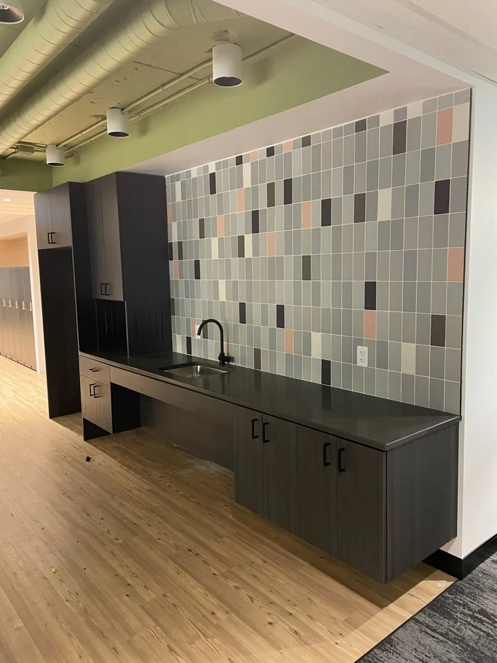 A kitchenette with gray cabinets, a black countertop, a black sink with a black faucet, a colorful tiled backsplash, and light wood flooring in an interior space with exposed ductwork and white ceiling.