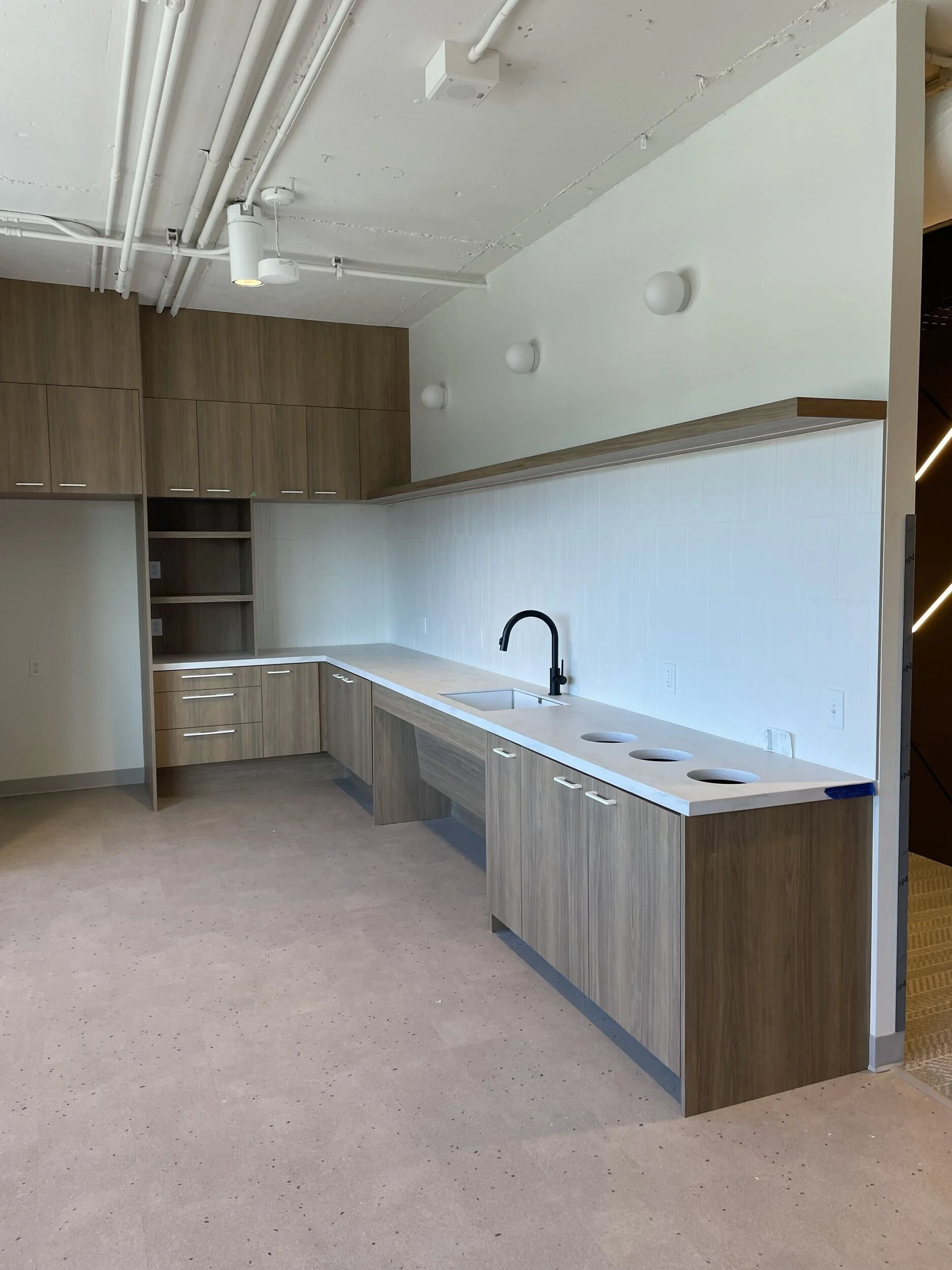 A modern kitchen under construction with wooden cabinets, a white countertop, and a black kitchen faucet. The ceiling has exposed pipes and light fixtures. The floor is unfinished, and the space appears to be in the process of being remodeled.