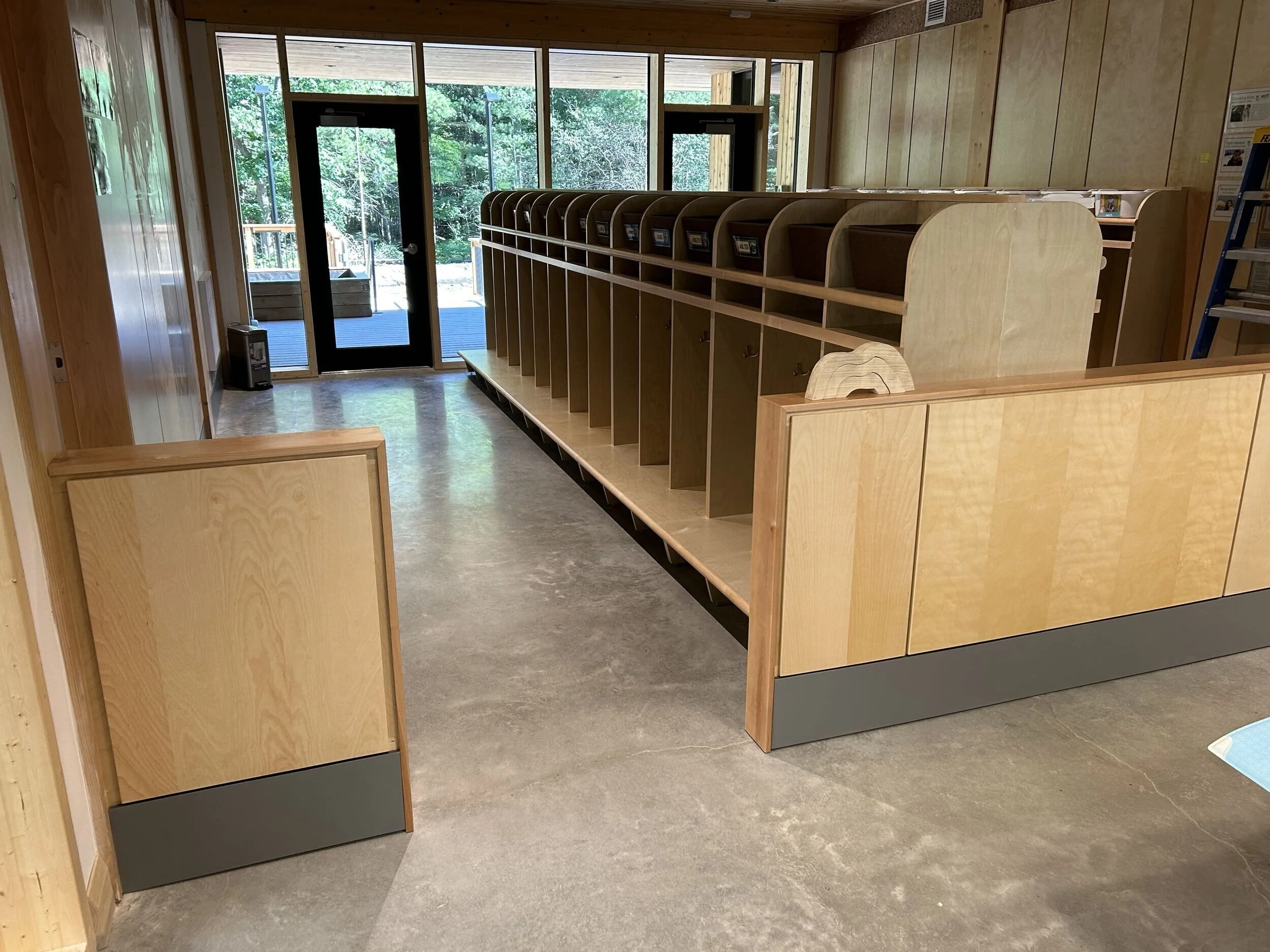 Interior view of a wooden public space with a row of coat hooks and cubbies, glass doors leading outside to a green wooded area.