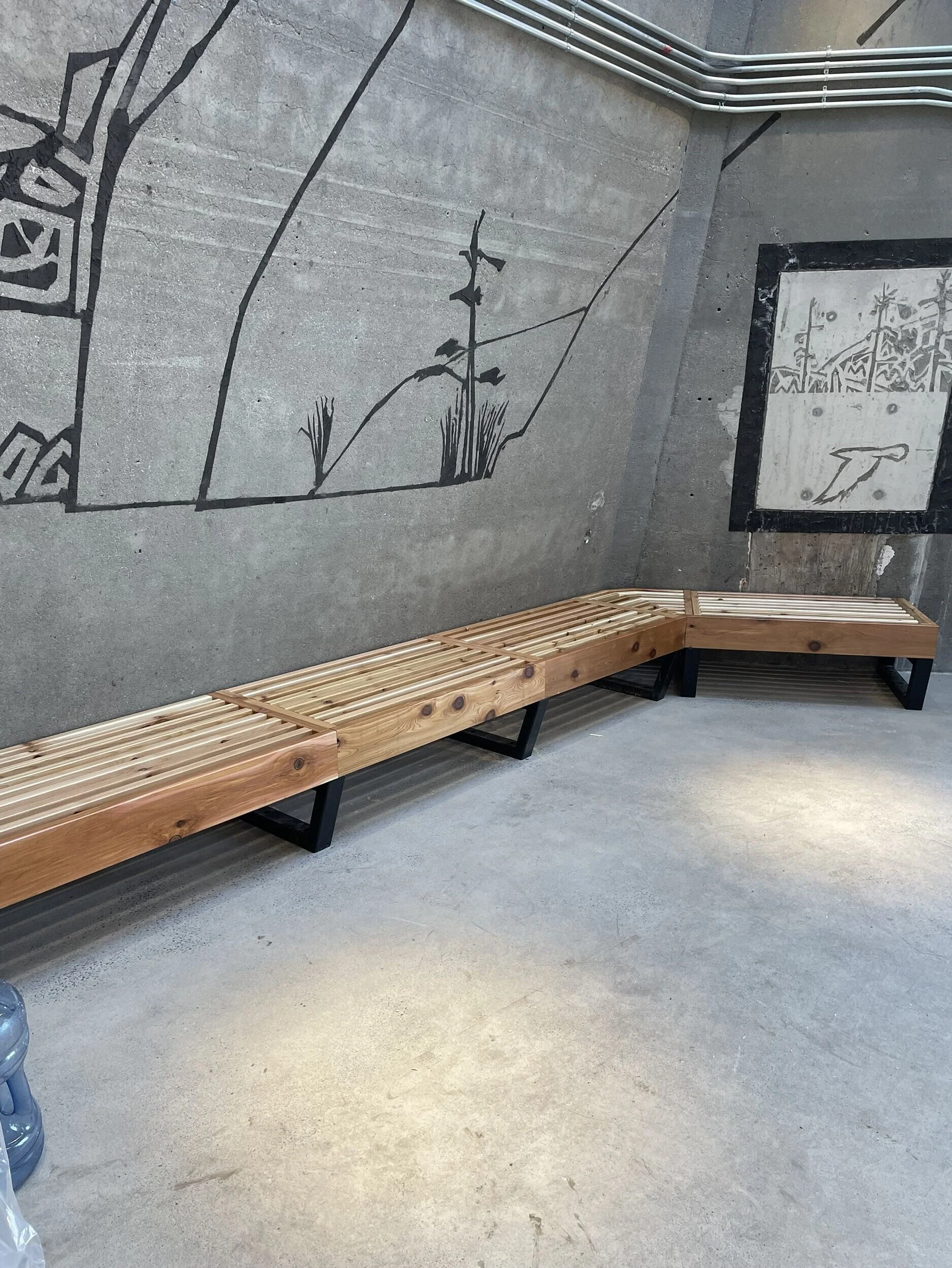 A metal wall with black outline artwork of a stem with leaves and grass. A black metal and wood bench in the foreground along the wall. A section of black-marble framed artwork on the right wall.