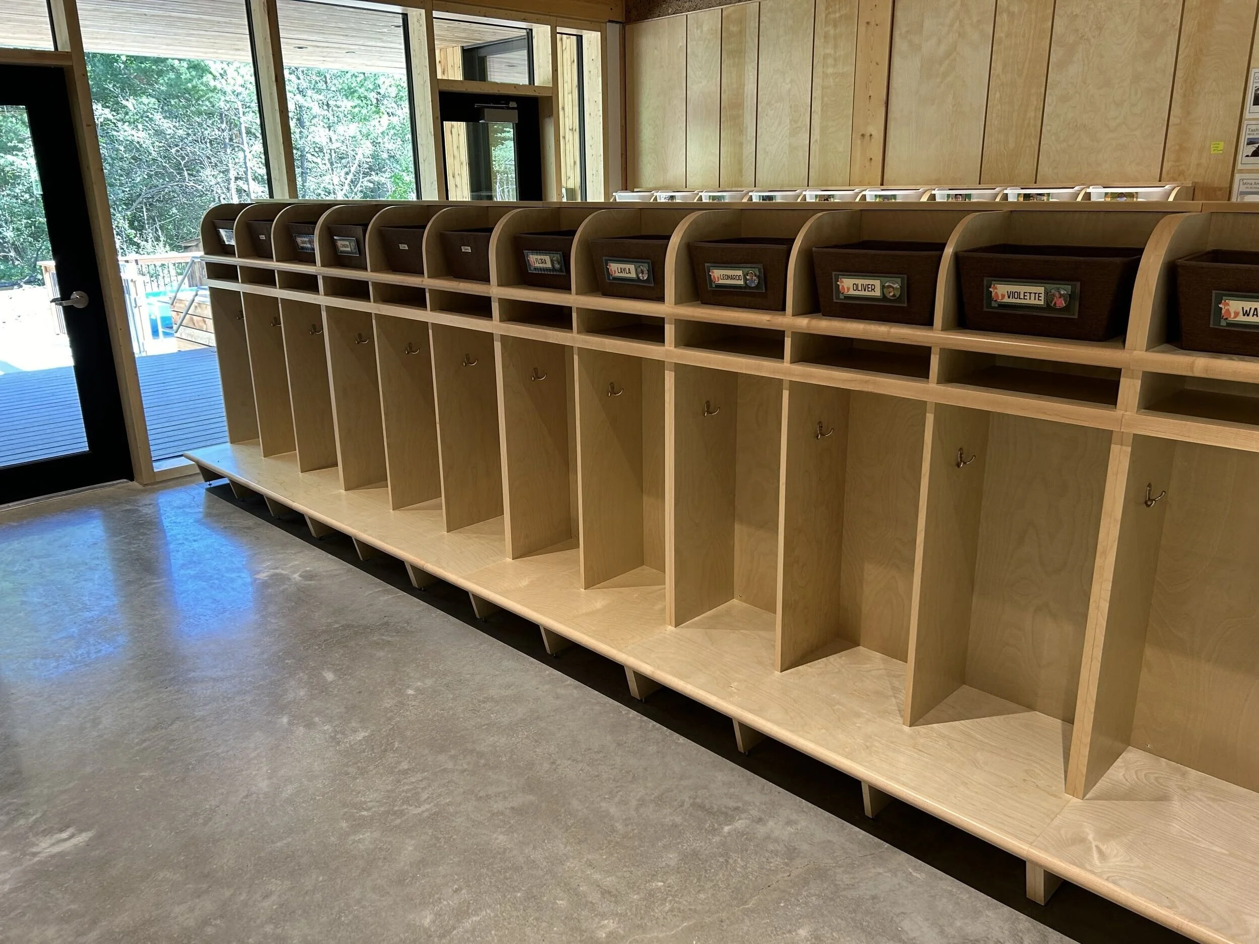 A wooden cubby rack with individual labeled compartments, each containing a basket, positioned against a wall in a room with large windows showing green trees outside.