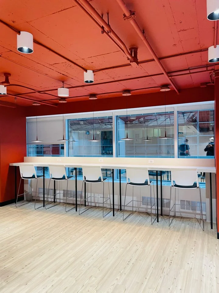 A bright modern seating area with a long white counter and six white bar stools near a large window, with a red ceiling and wooden floor.