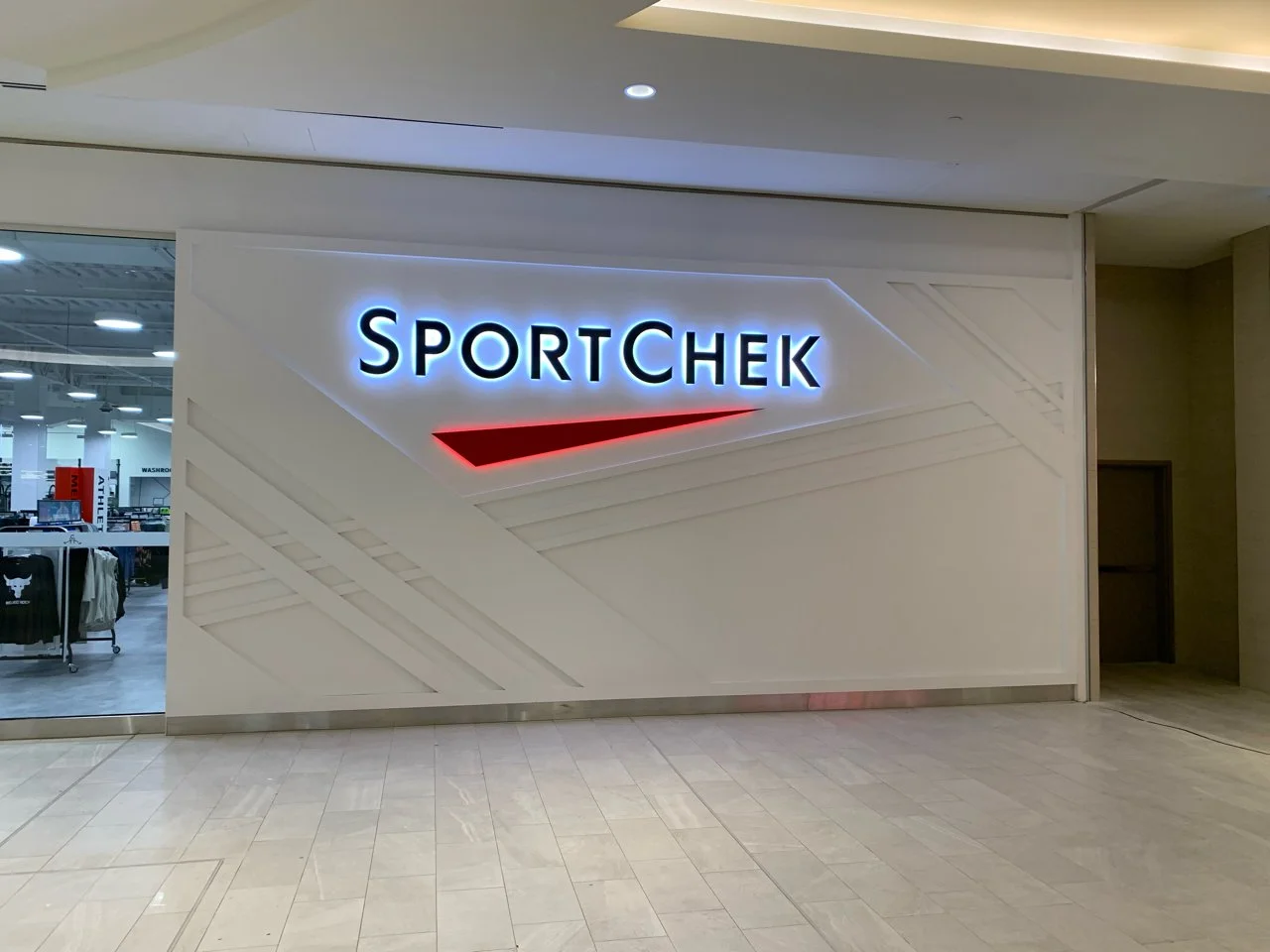 Sign that reads 'SPORTCHEK' in illuminated letters with a red and black swoosh logo below it, outside a retail store.