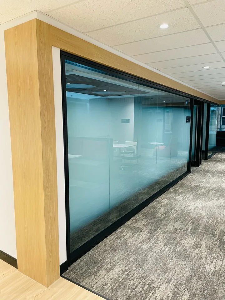 Glass office or conference room with wooden framing and carpeted floor, seen from the hallway in a modern building.