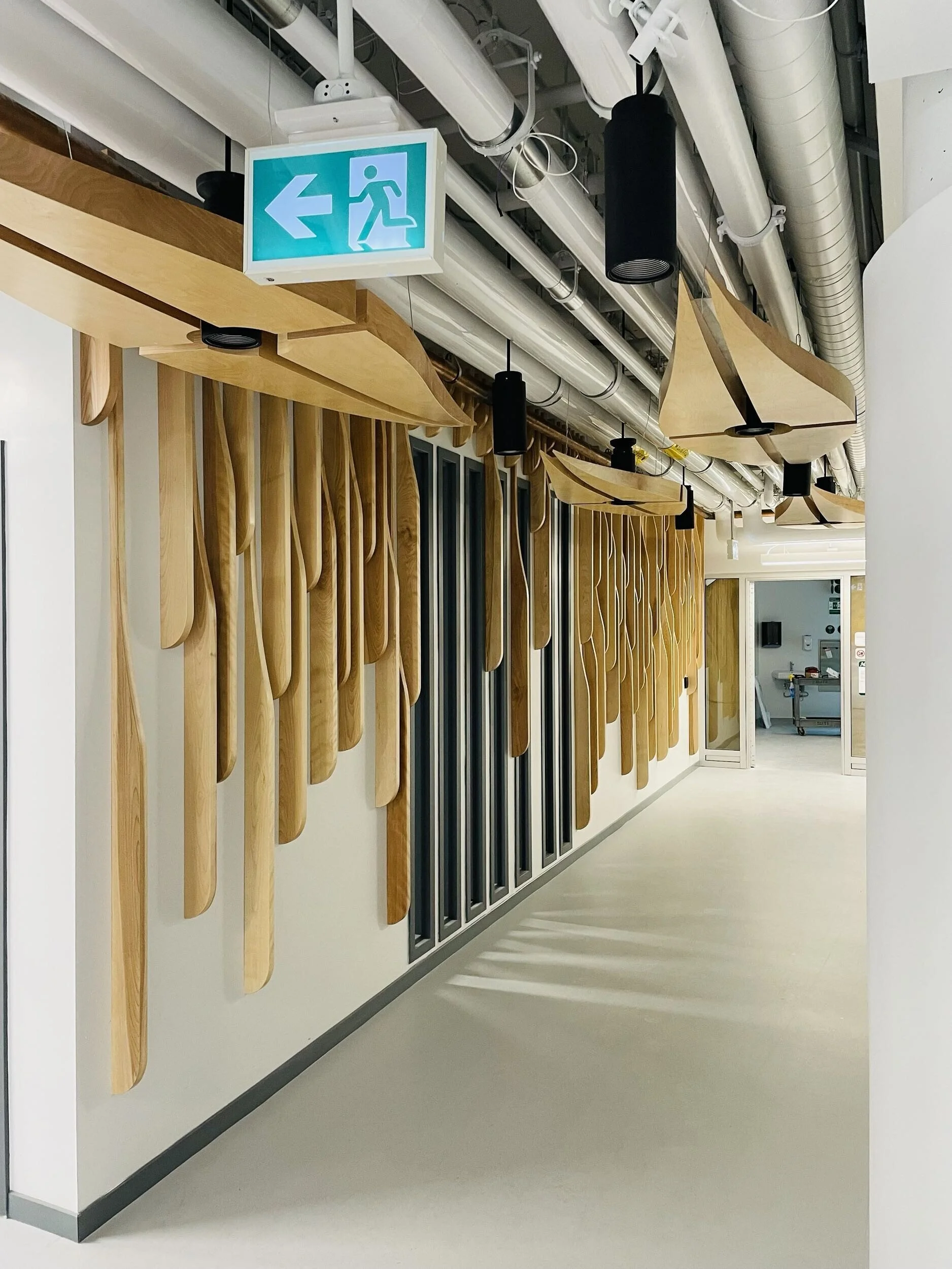 A ceiling with metal ducts and black cylindrical light fixtures, a wooden decorative panel with skateboard shapes, and an emergency exit sign pointing to the left.