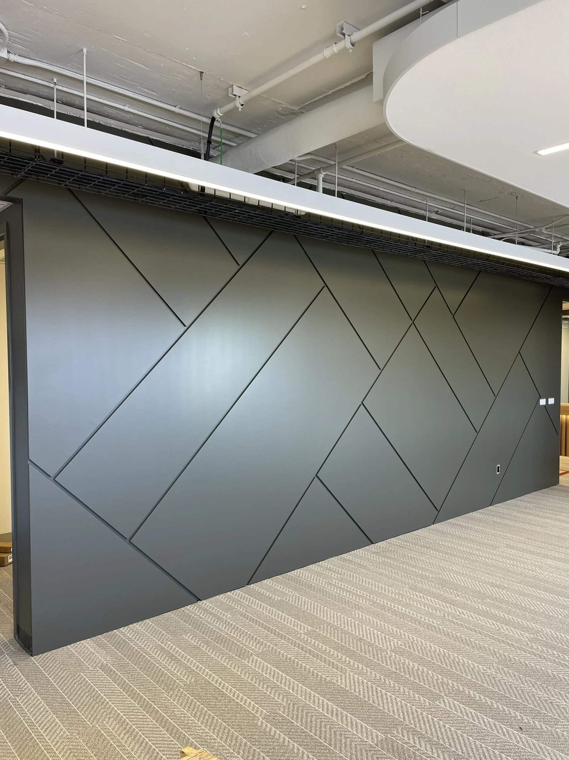 Modern interior space with a grey geometric partition wall, suspended white ceiling, and visible industrial piping, carpeted flooring with a striped pattern.