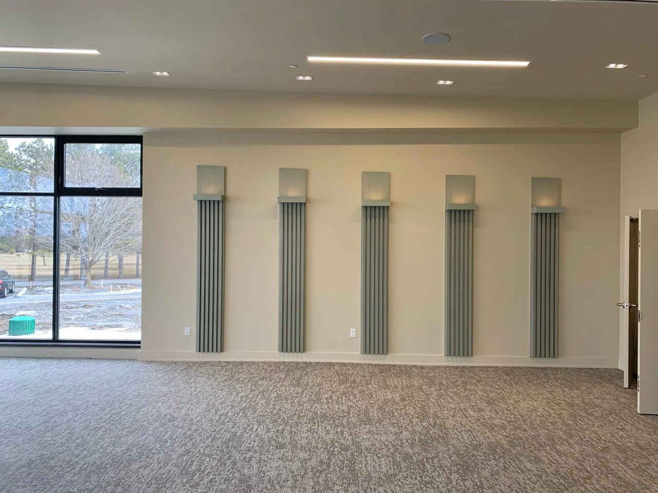 Empty room with five vertical sound diffusers on the wall, large window revealing trees and cars outside, beige carpet, and a door on the right.