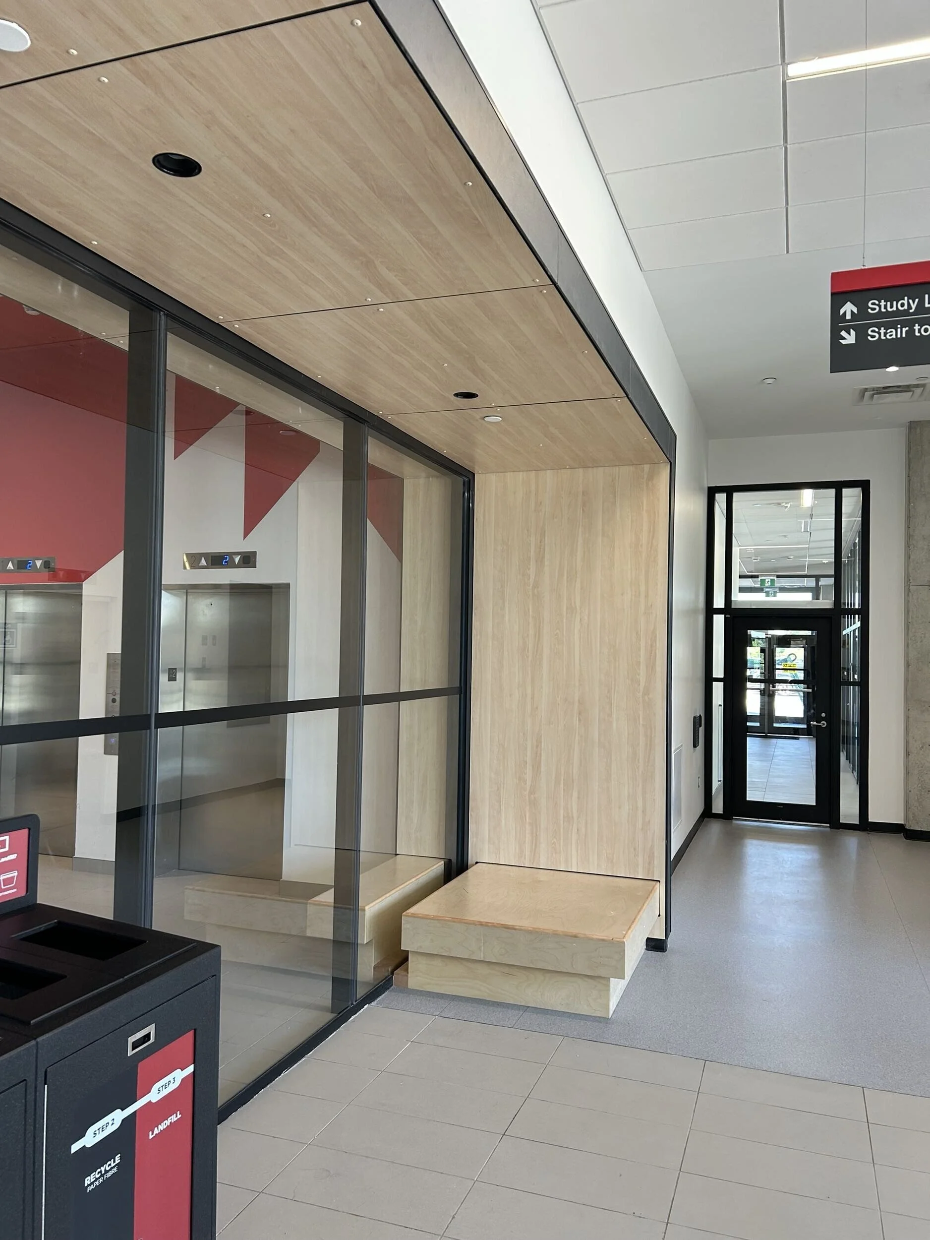 Interior of a modern building lobby with elevators, a wood-paneled ceiling, a digital directory sign, and a black recycling bin.