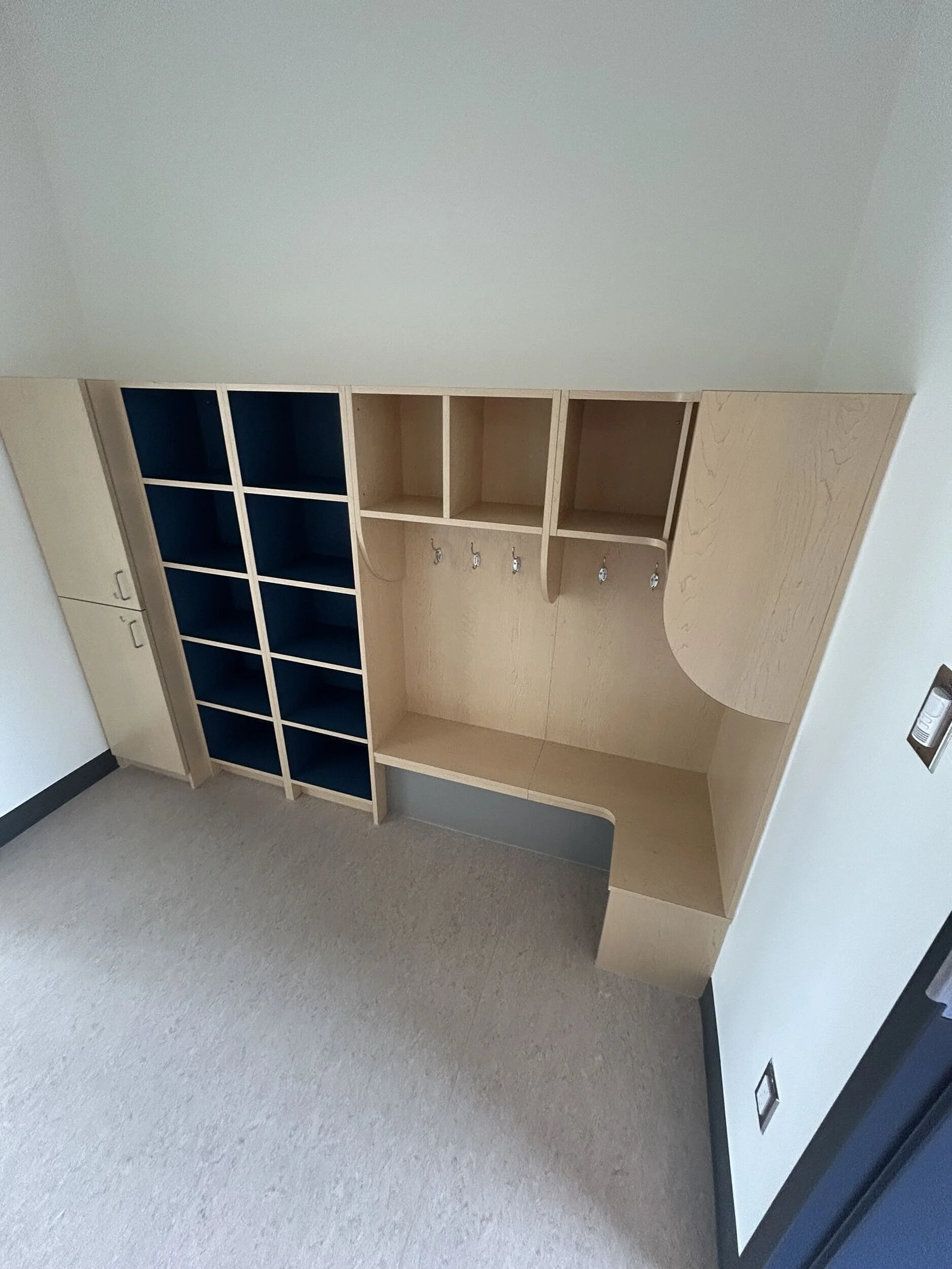Empty wooden cubicle storage unit with black cubicles on the left, hooks underneath middle shelf, and a curved desk on the right side, against a white wall and beige carpeted floor.
