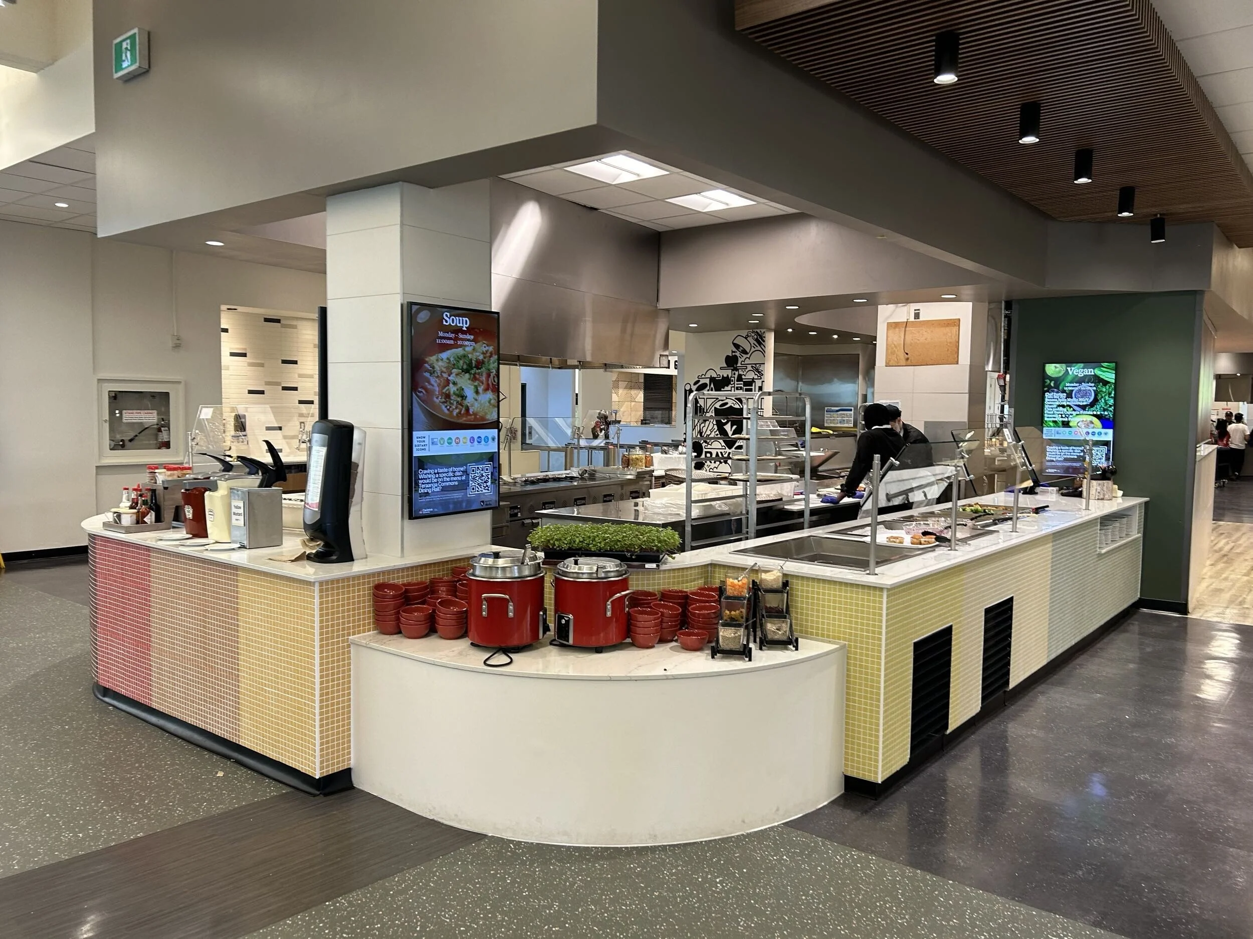 A food court counter with digital menu screens displaying soup and vegan options. The counter has red bowls, condiments, and a food warmer. The background shows part of a kitchen and restaurant area with staff preparing food.