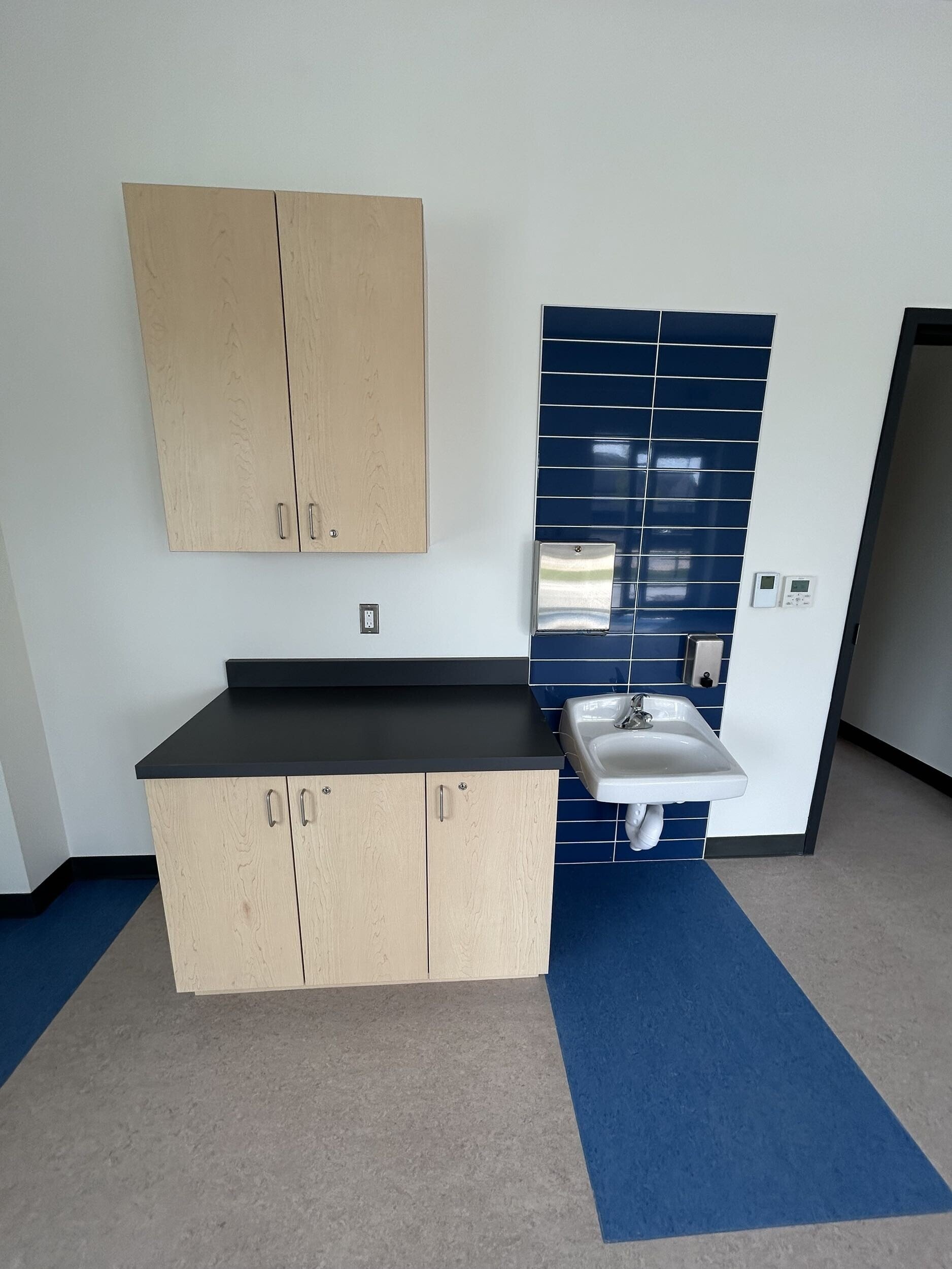 Interior of a room with a white wall, beige cabinets, a blue tiled wall, a sink, and a dark counter.
