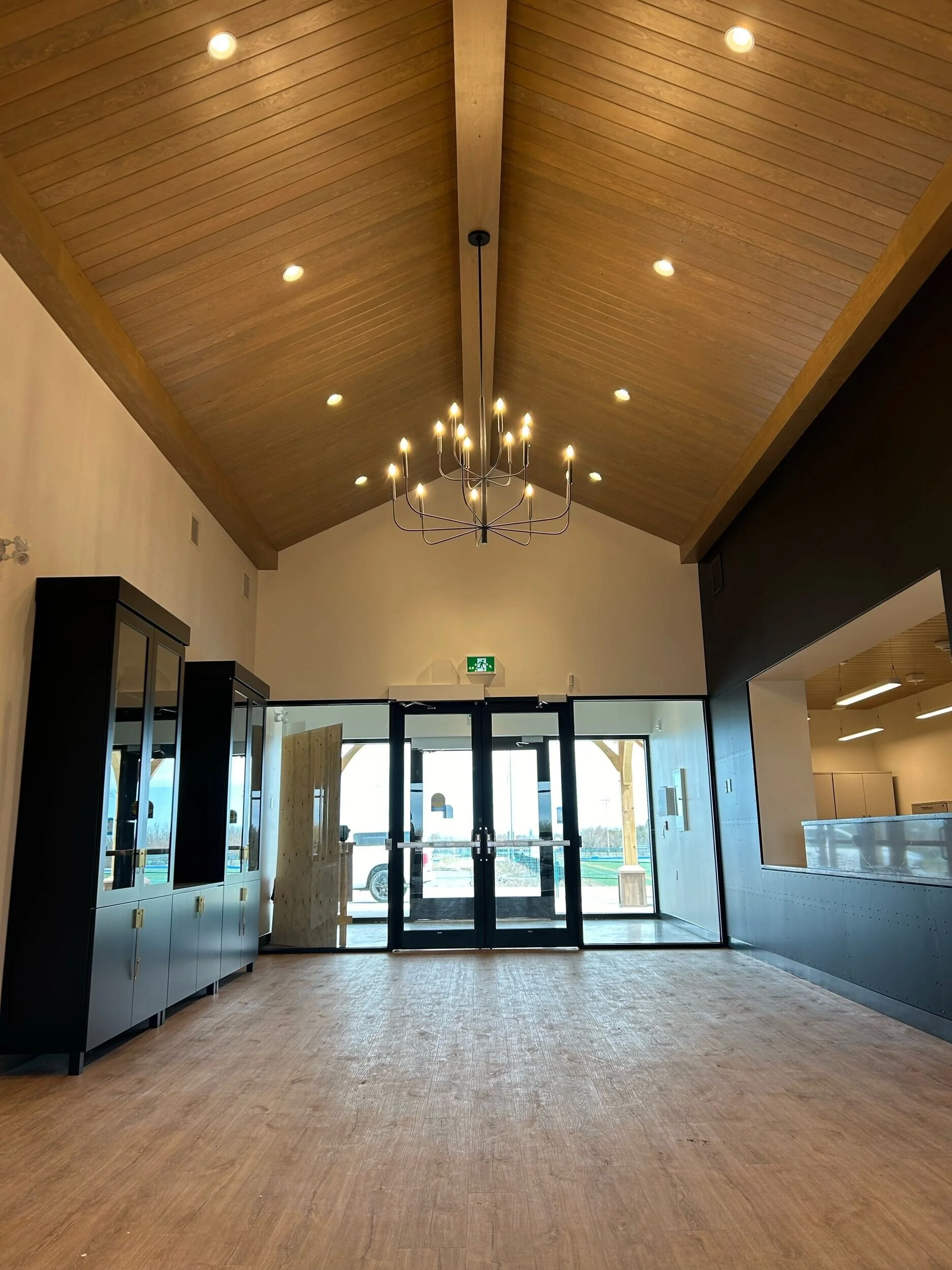 Interior view of a modern building with a wooden vaulted ceiling, chandelier, glass double doors, and light flooring.