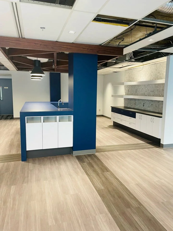 Interior of a modern office or commercial space with a kitchenette area featuring a blue and white color scheme, open ceiling with exposed panels and lighting, and light wood flooring.