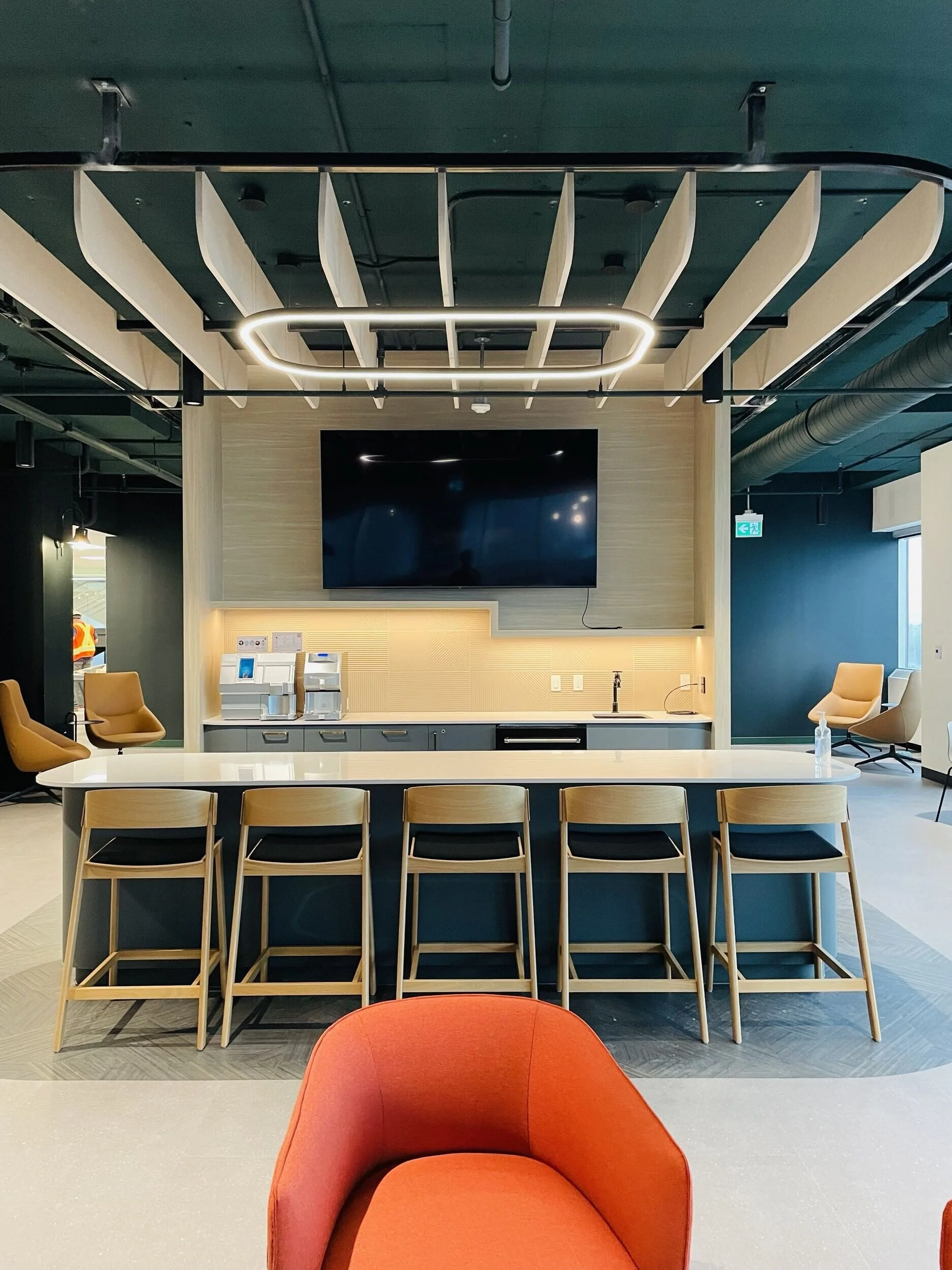 Modern office lounge with a large conference table, orange chairs, and a high-top with four bar stools in front of a kitchenette area. A large flat-screen TV is mounted on the wall above the kitchenette. The ceiling features artistic wooden slats and