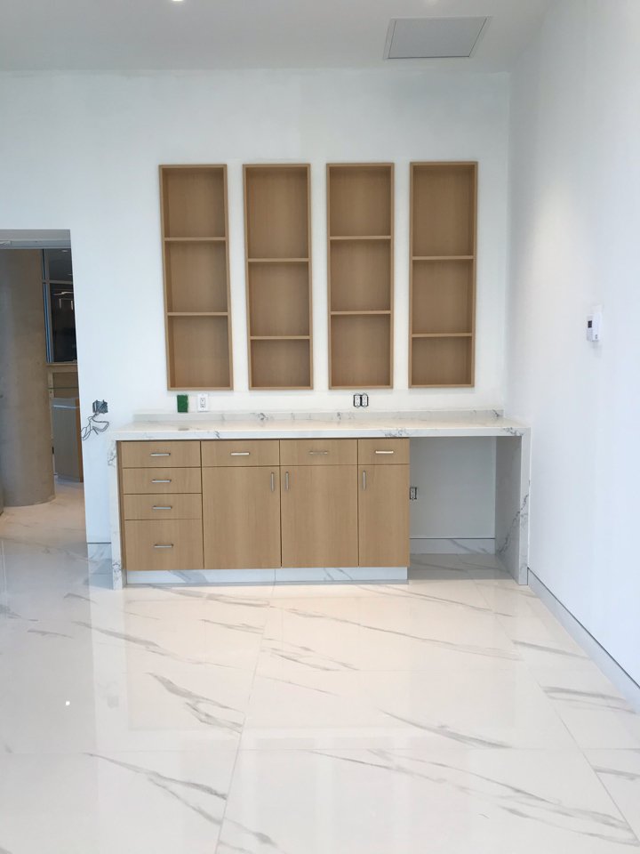 Empty kitchen cabinet with four tall open shelves above a countertop, which has a lower set of cabinets and drawers, all made of light-colored wood. The floor is shiny with a marble pattern, and the wall behind is white.