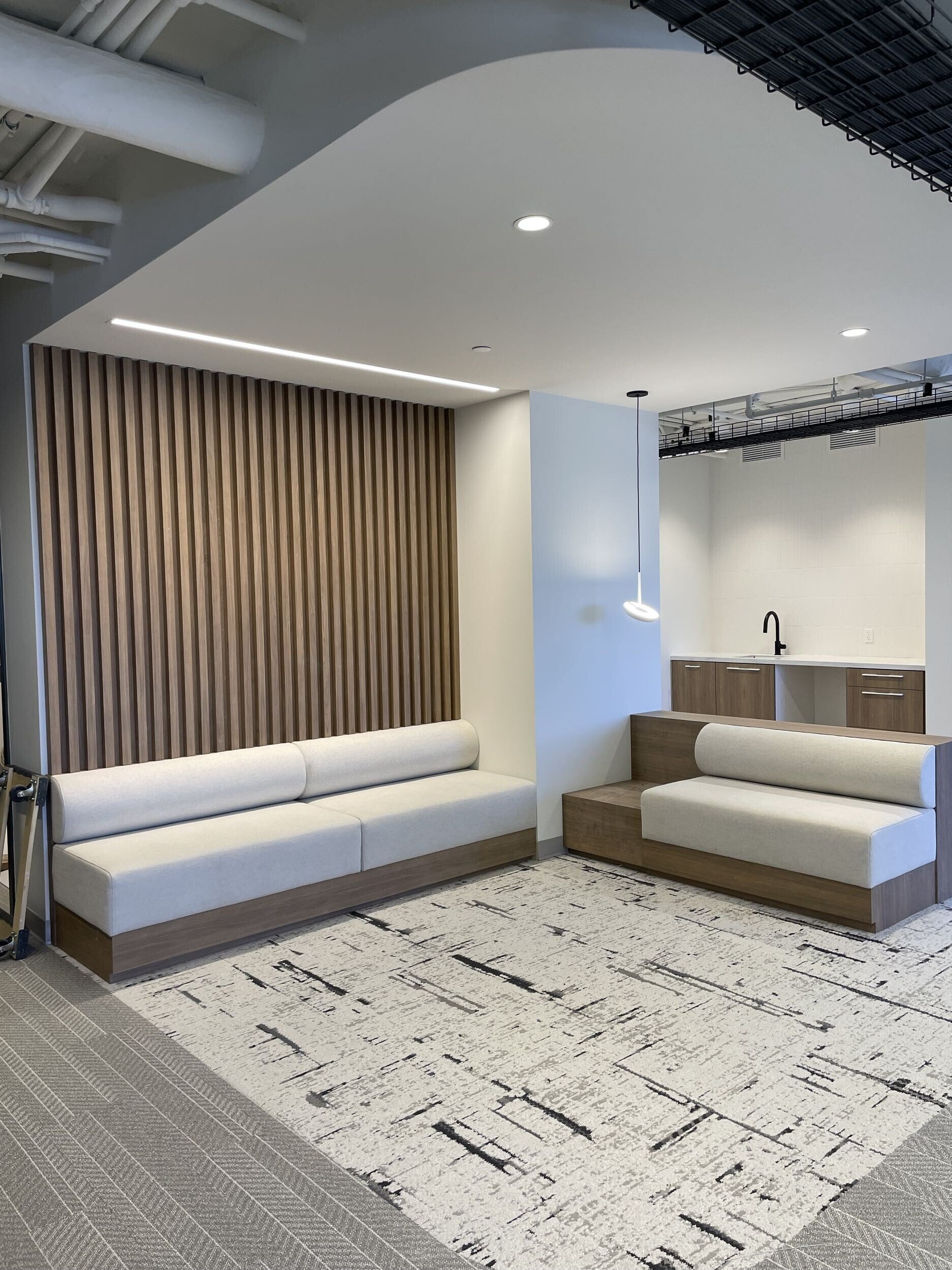 Modern office or lounge area with two white cushioned sofas with wooden bases, a textured patterned rug, and a wooden slatted wall panel. The space has neutral colors, a sleek ceiling with recessed and hanging lights, exposed ductwork, and an open, i