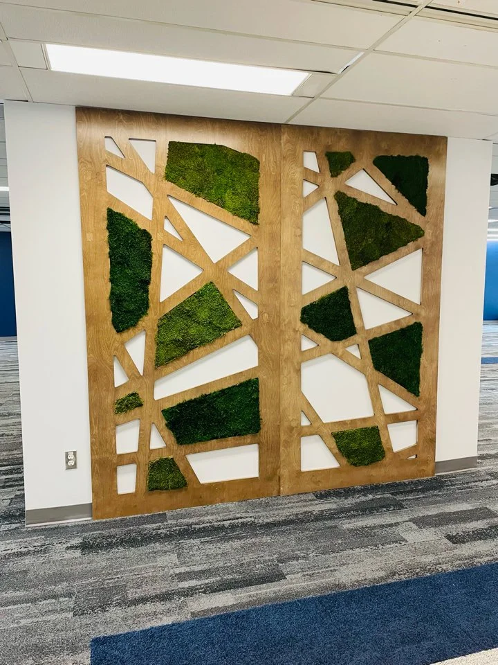 Decorative wooden wall panel with geometric cutouts filled with green moss, located in an office corridor.